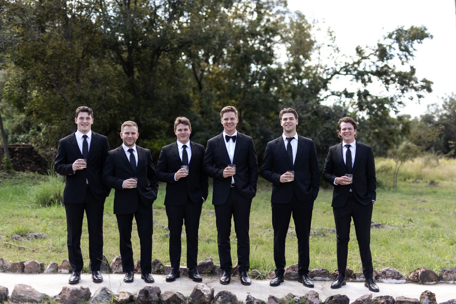 Groomsmen in black suits standing in line on stone wall outdoors.