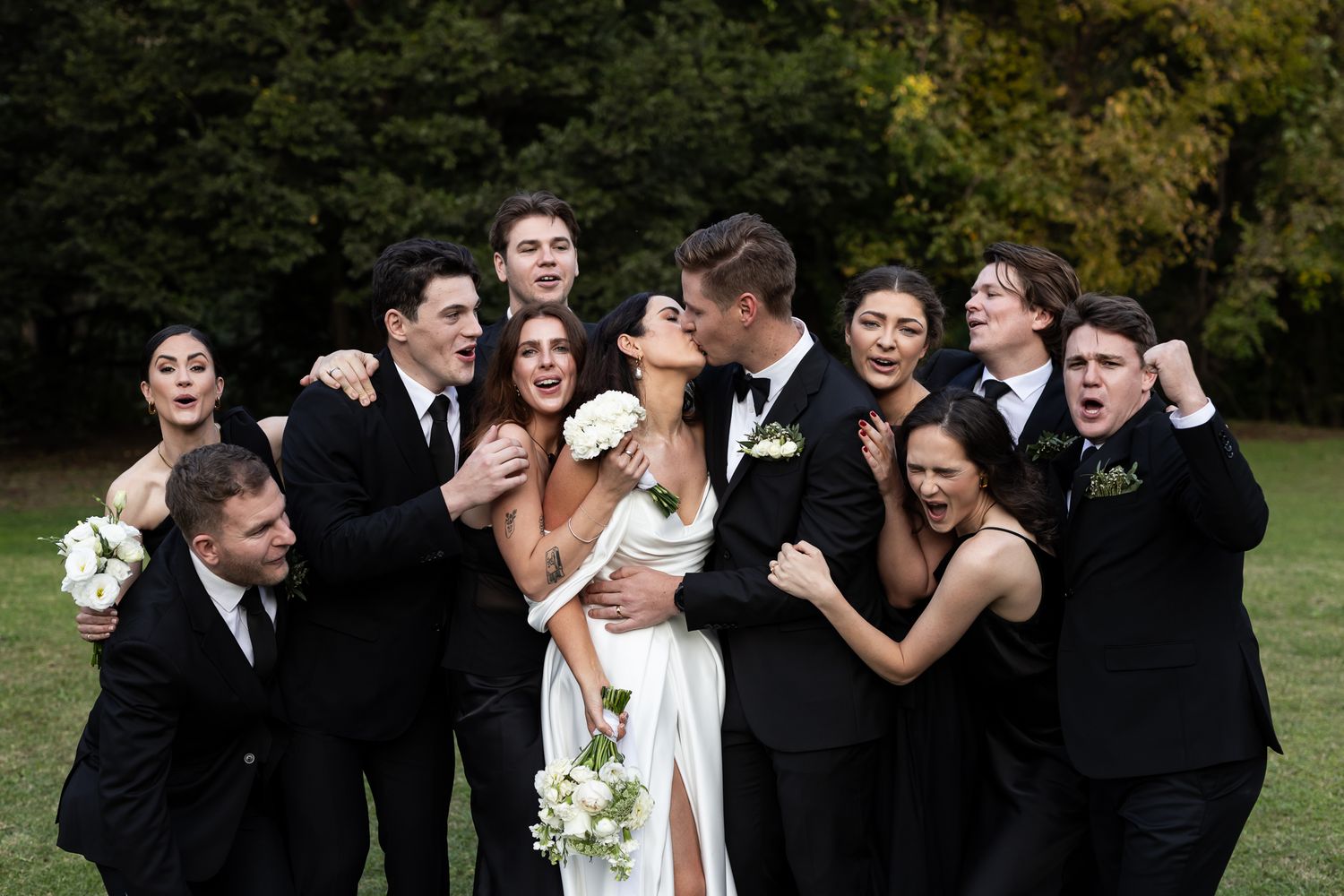 A joyful wedding party shares an energetic group moment on grass with trees in the background.