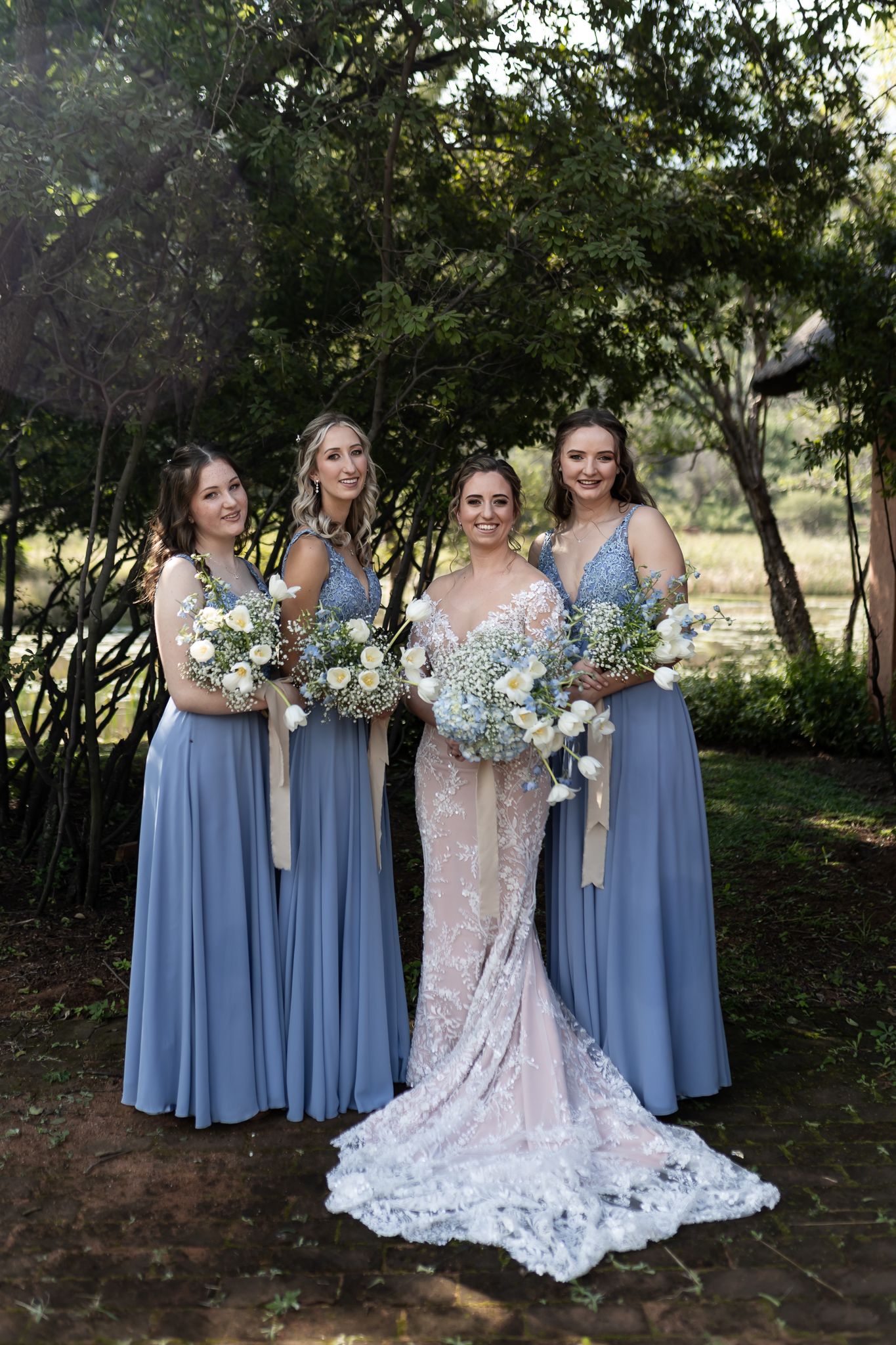 A wedding party in dusty blue dresses and lace gown pose together outdoors with floral bouquets.