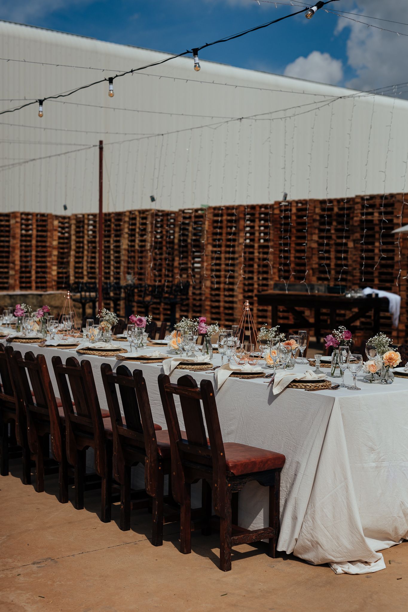 Outdoor wedding venue with long table setup against rustic wooden wall and string lights.