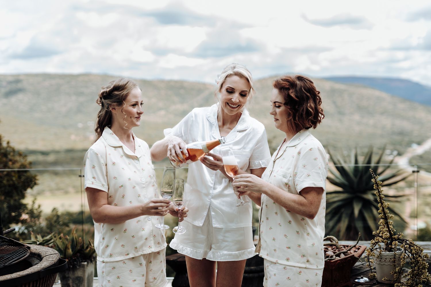 Three women in white robes toasting on luxurious mountain deck during celebration.
