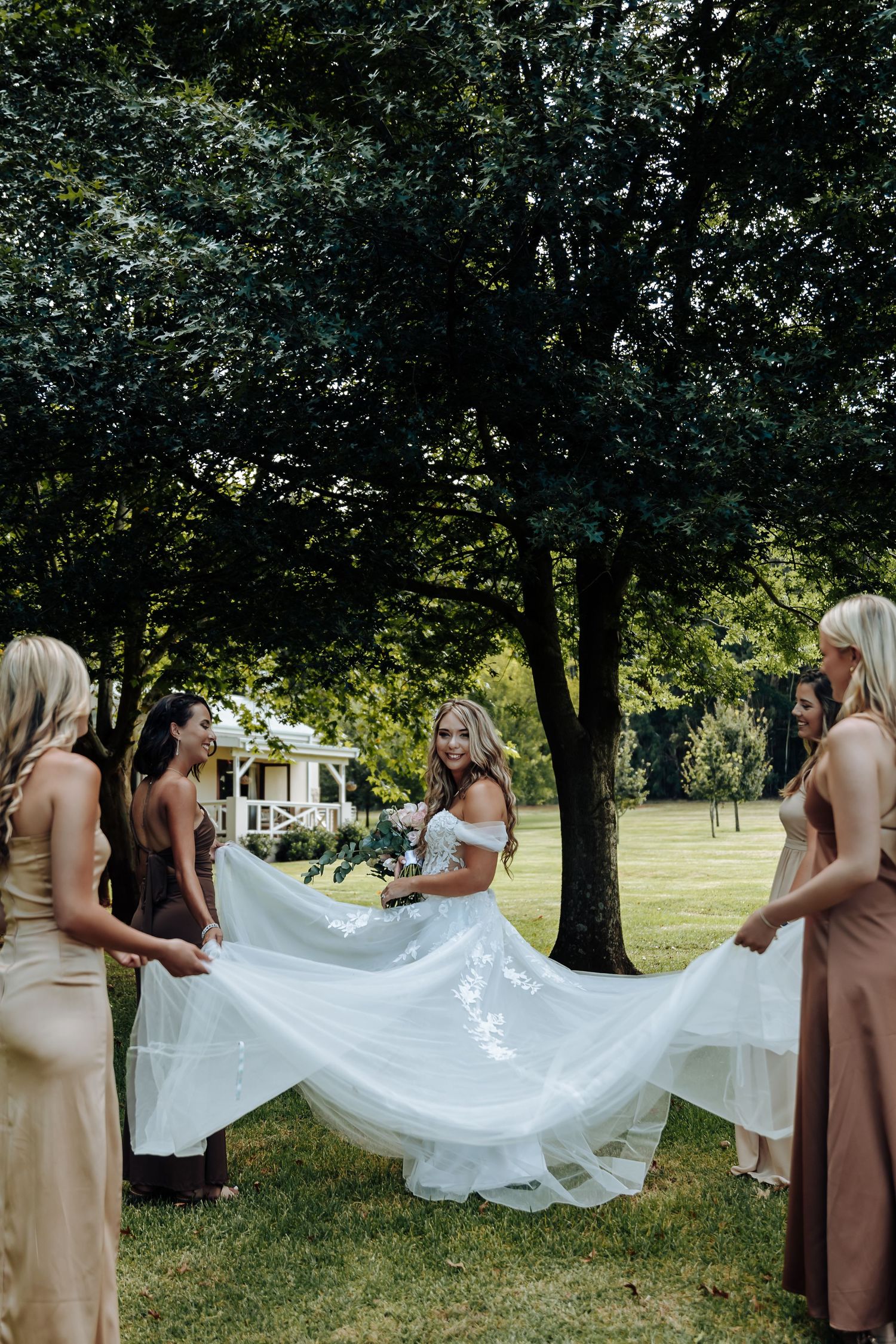 Bridesmaids in beige dresses help arrange the bride's flowing white veil on a grassy lawn surrounded by trees.