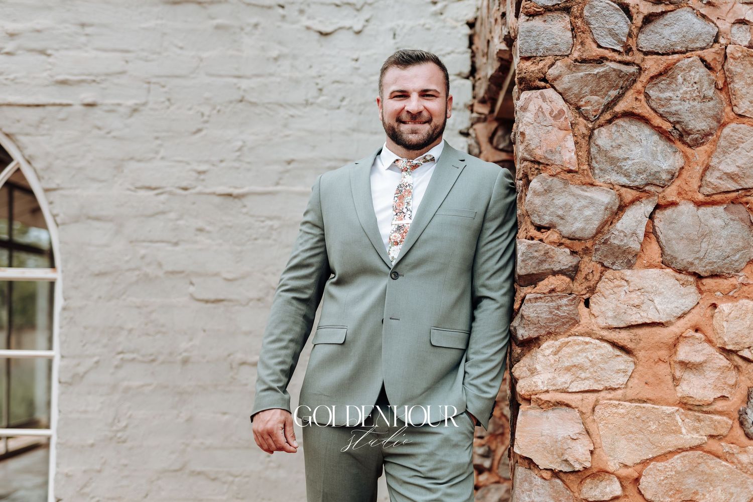 Person in a light sage green suit poses against a rustic stone wall background.