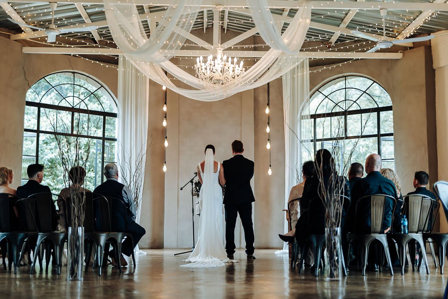 A wedding ceremony in a bright venue with arched windows, white drapes, a chandelier and guests seated in grey chairs.