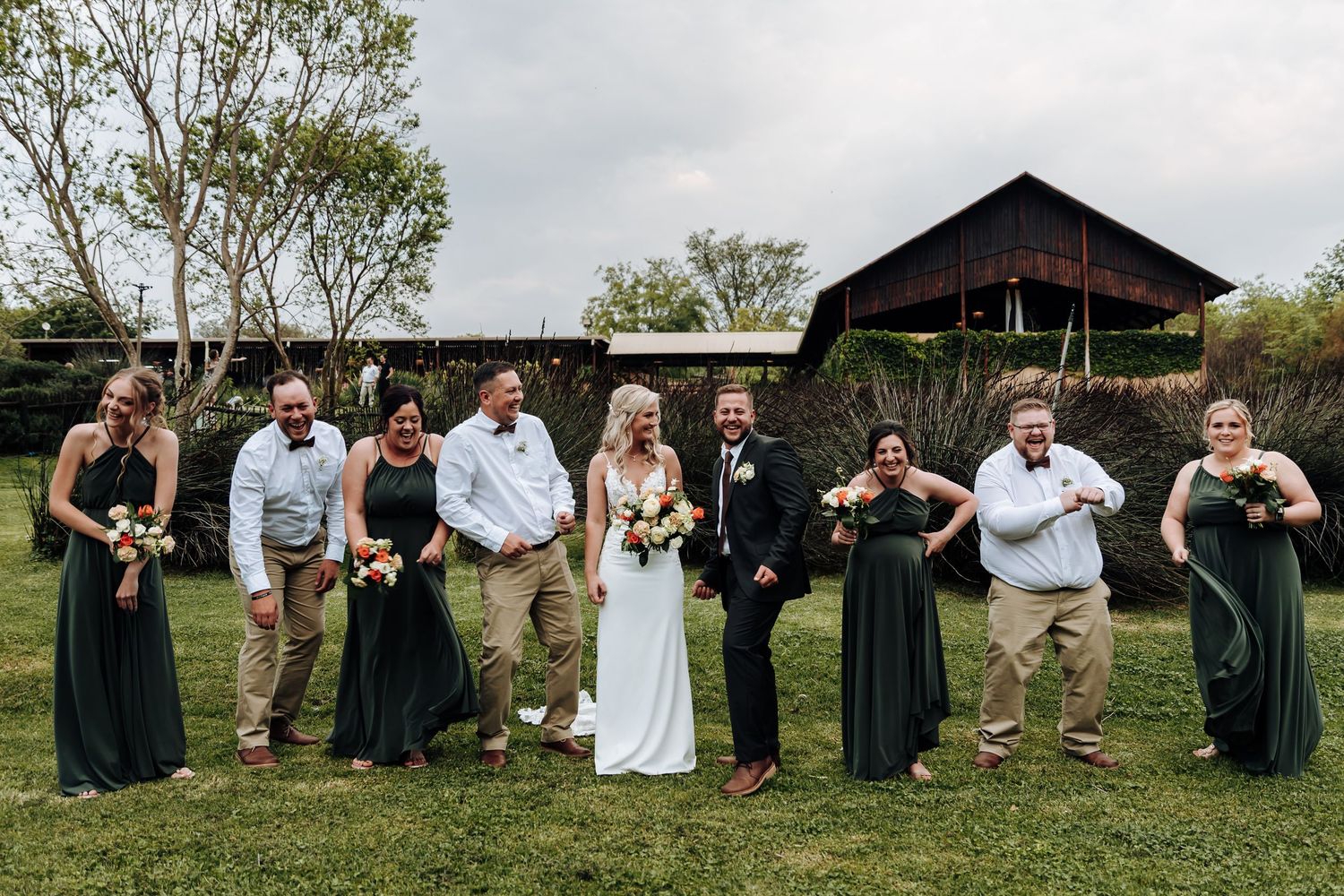Wedding party members stand in a row against a rustic barn backdrop during an outdoor celebration.