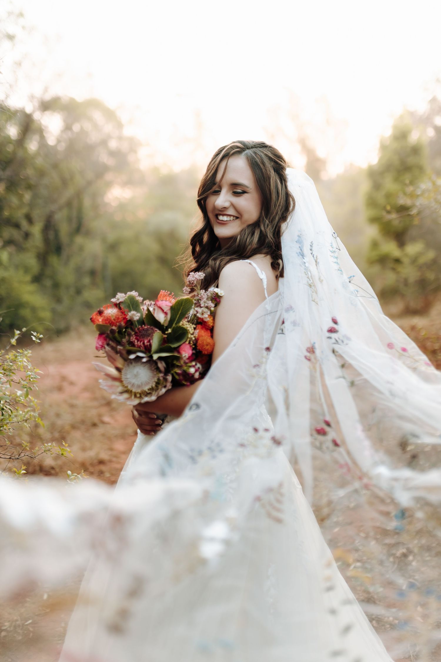 A flowing white wedding veil catches the breeze as a bride poses with a vibrant red and orange bouquet at sunset.