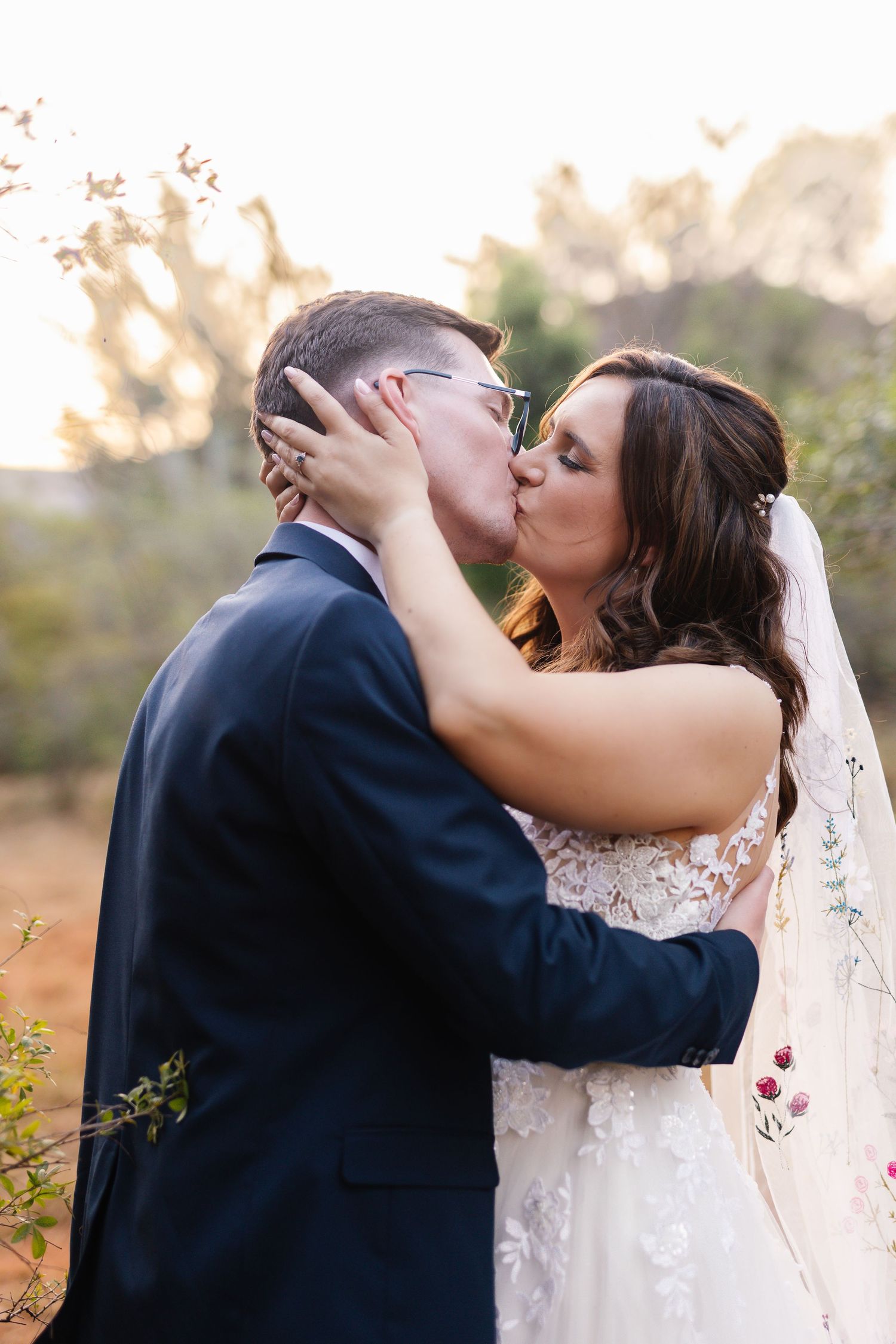 A romantic wedding couple embraces and kisses in a beautiful outdoor setting during golden hour.
