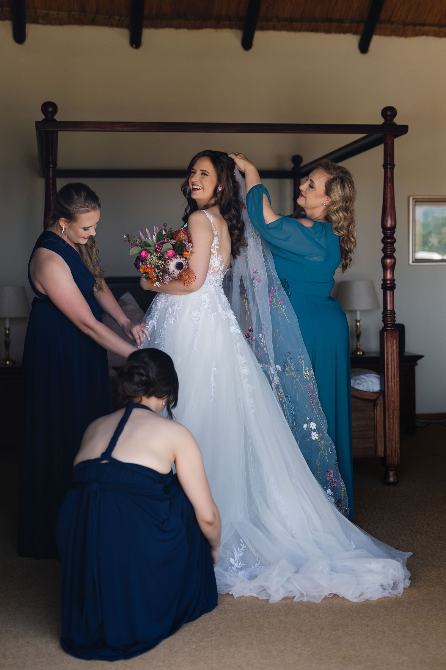 A bride in a white lace wedding gown with flowing veil gets help from attendants in navy and blue dresses.