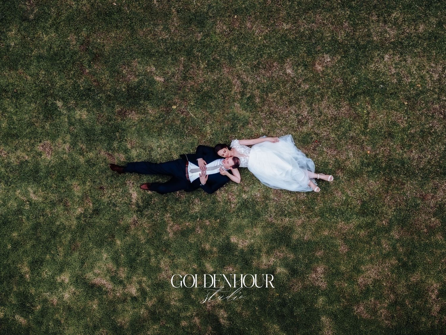 Aerial drone view of bride and groom lying on green grass in wedding attire.