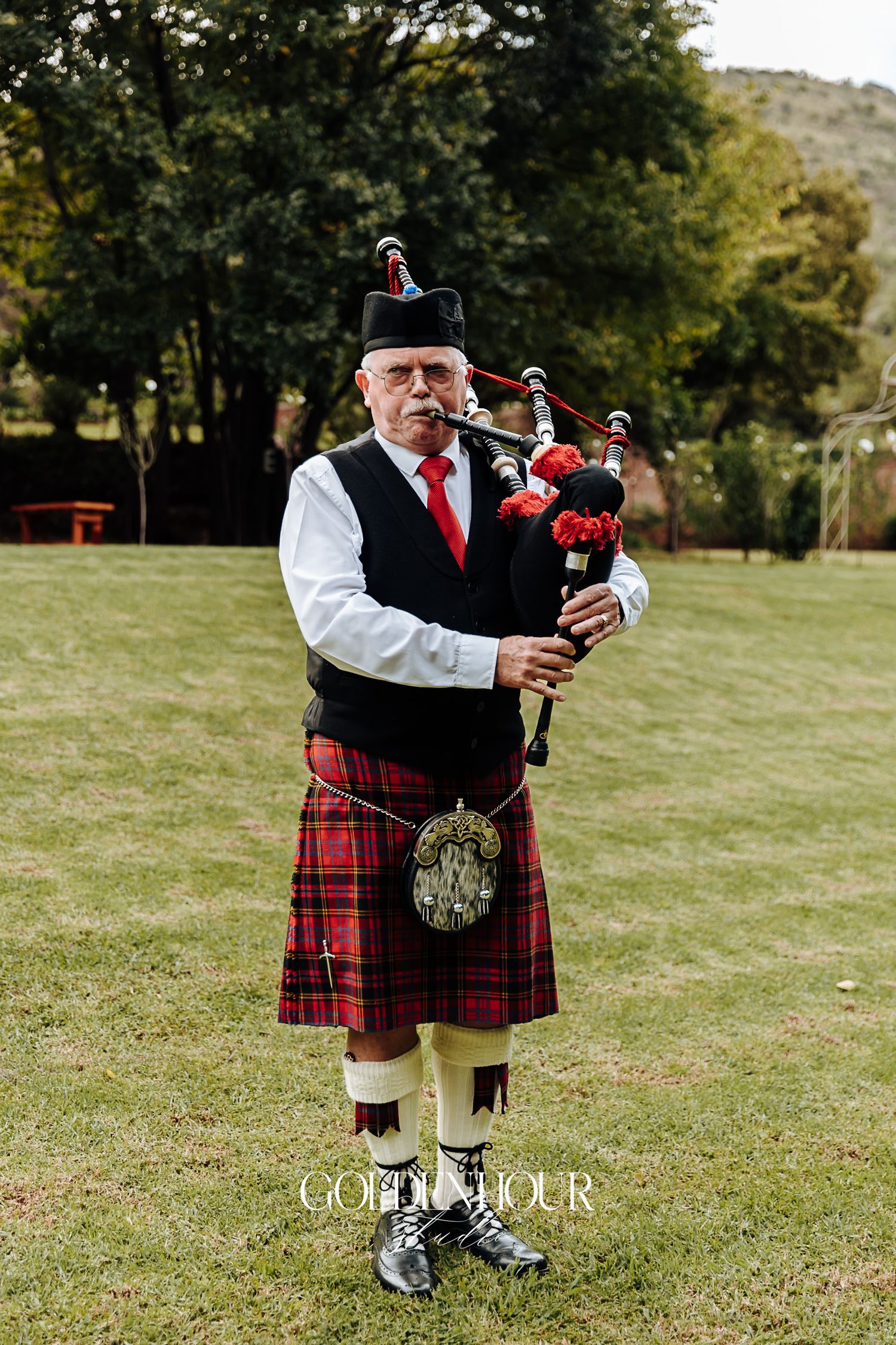 Traditional Scottish bagpiper in red tartan kilt and black vest performing on green lawn at wedding.