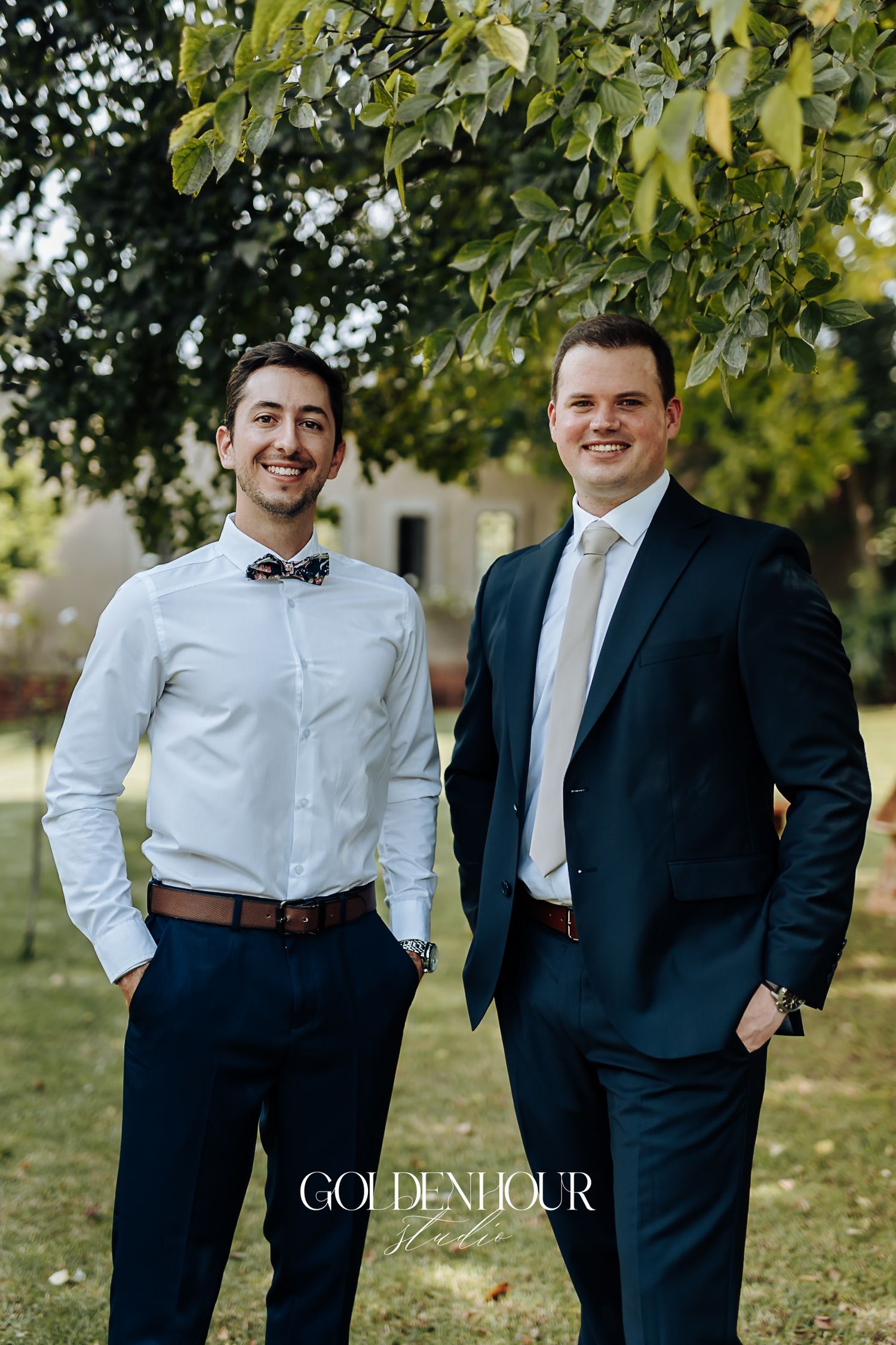 Two smiling groomsmen in navy suits posing outdoors under green trees at a wedding.