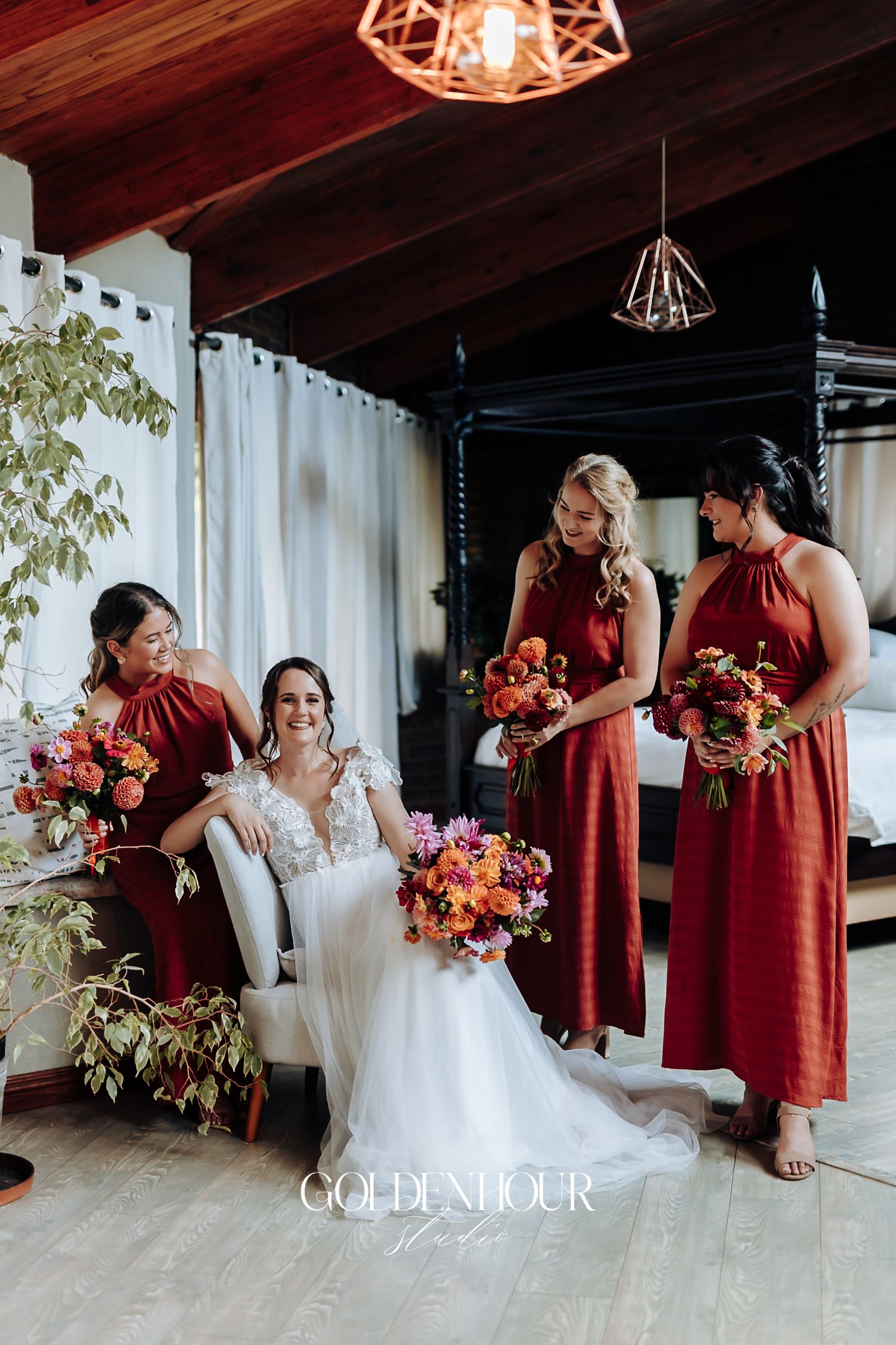Bride in lace gown seated with three bridesmaids in rust red dresses holding colorful bouquets.