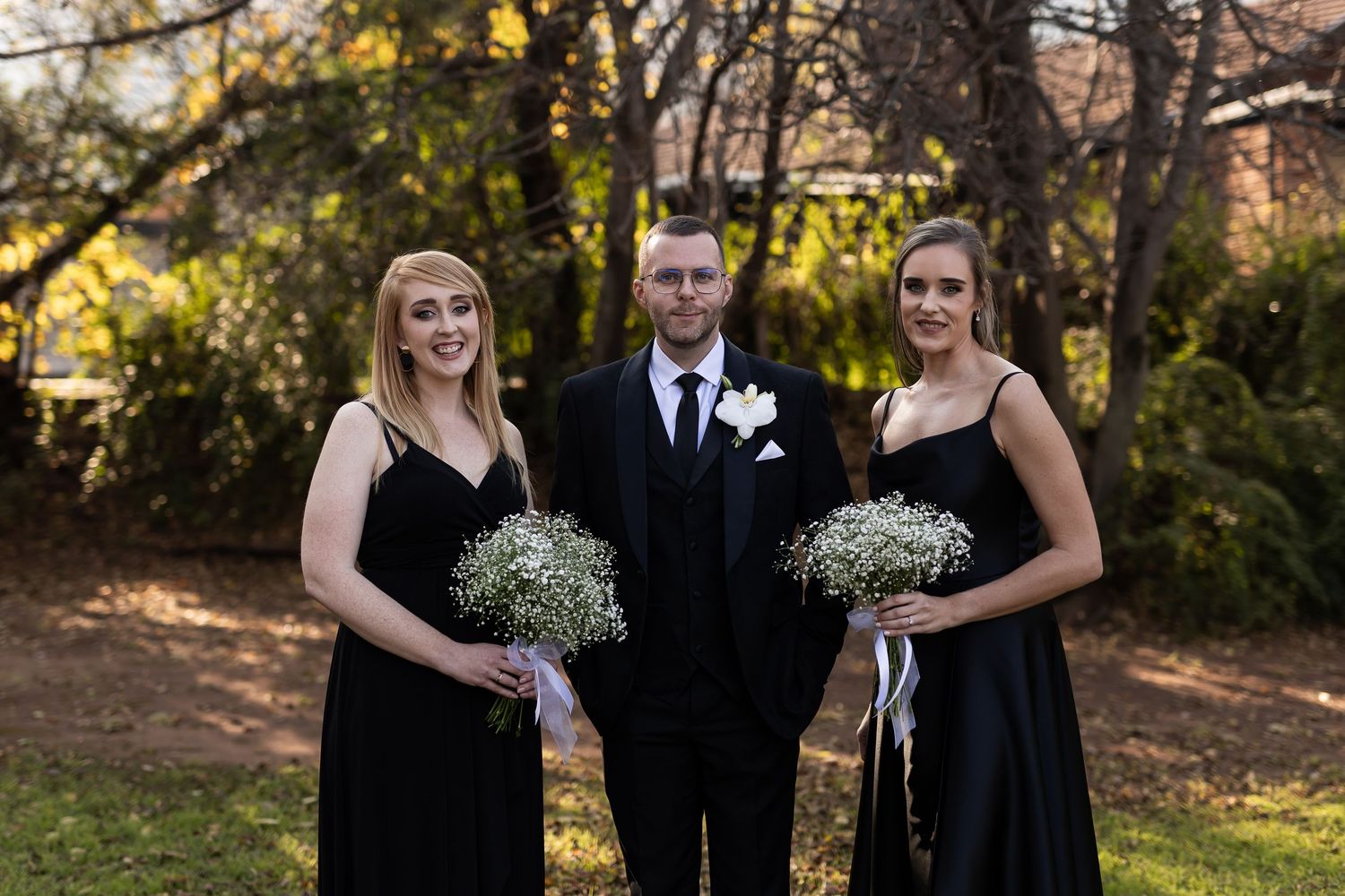 Wedding party members wearing elegant black attire stand together holding white baby's breath bouquets in an outdoor garden setting.