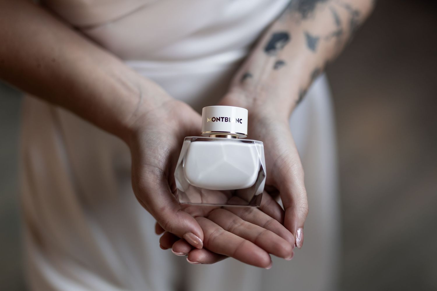 Tattooed hands gently hold a small white ceramic jar against a soft blurred background.