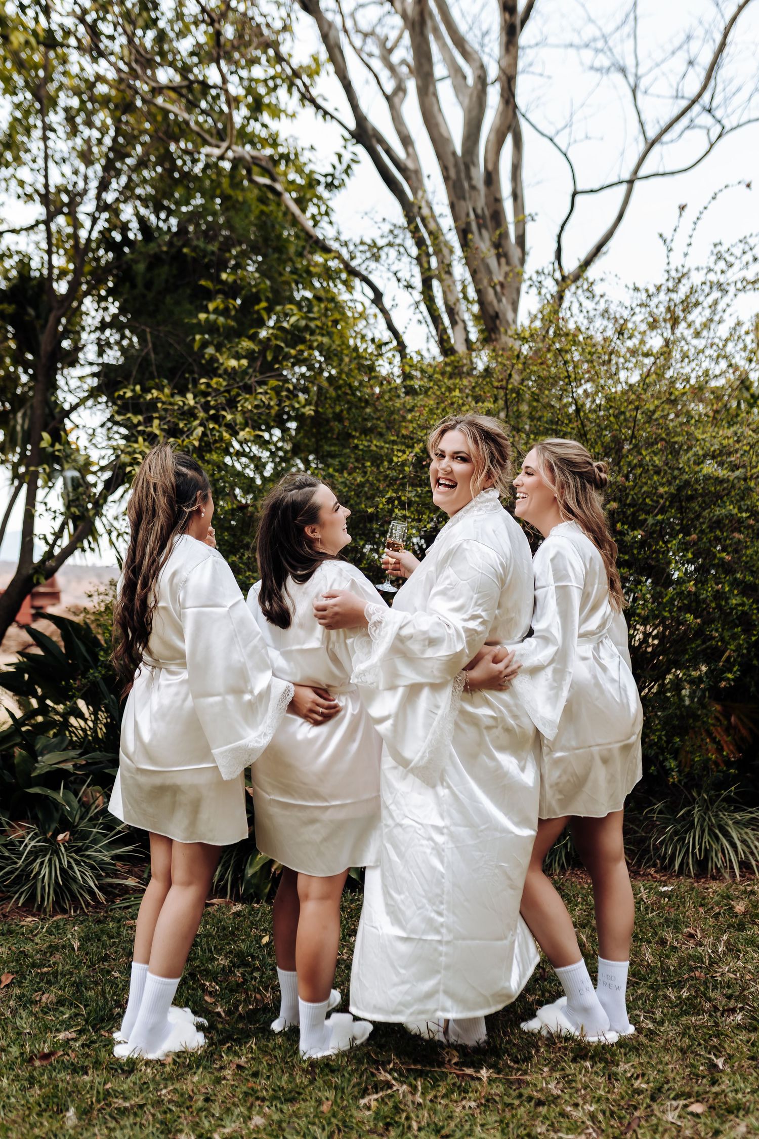 A group of friends wearing matching white outfits celebrate and dance together on a grassy hillside during a photoshoot.