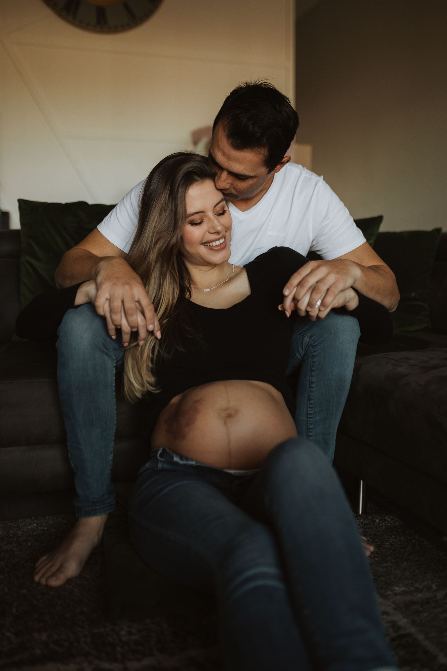 A couple snuggled together on a dark couch share an intimate maternity moment in casual denim clothing.