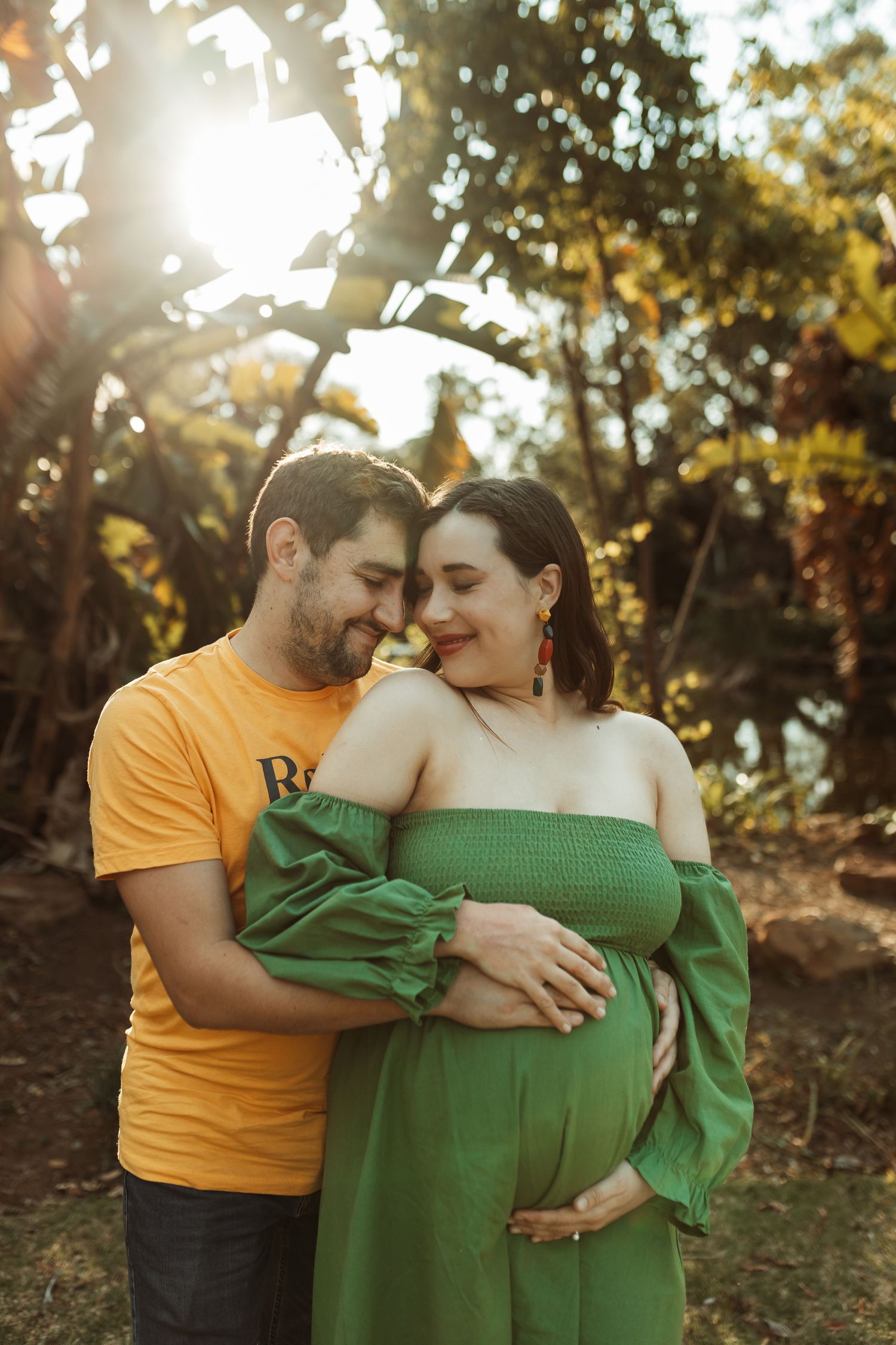 A couple embraces affectionately in warm sunlight, wearing yellow and green outfits in a wooded outdoor setting.