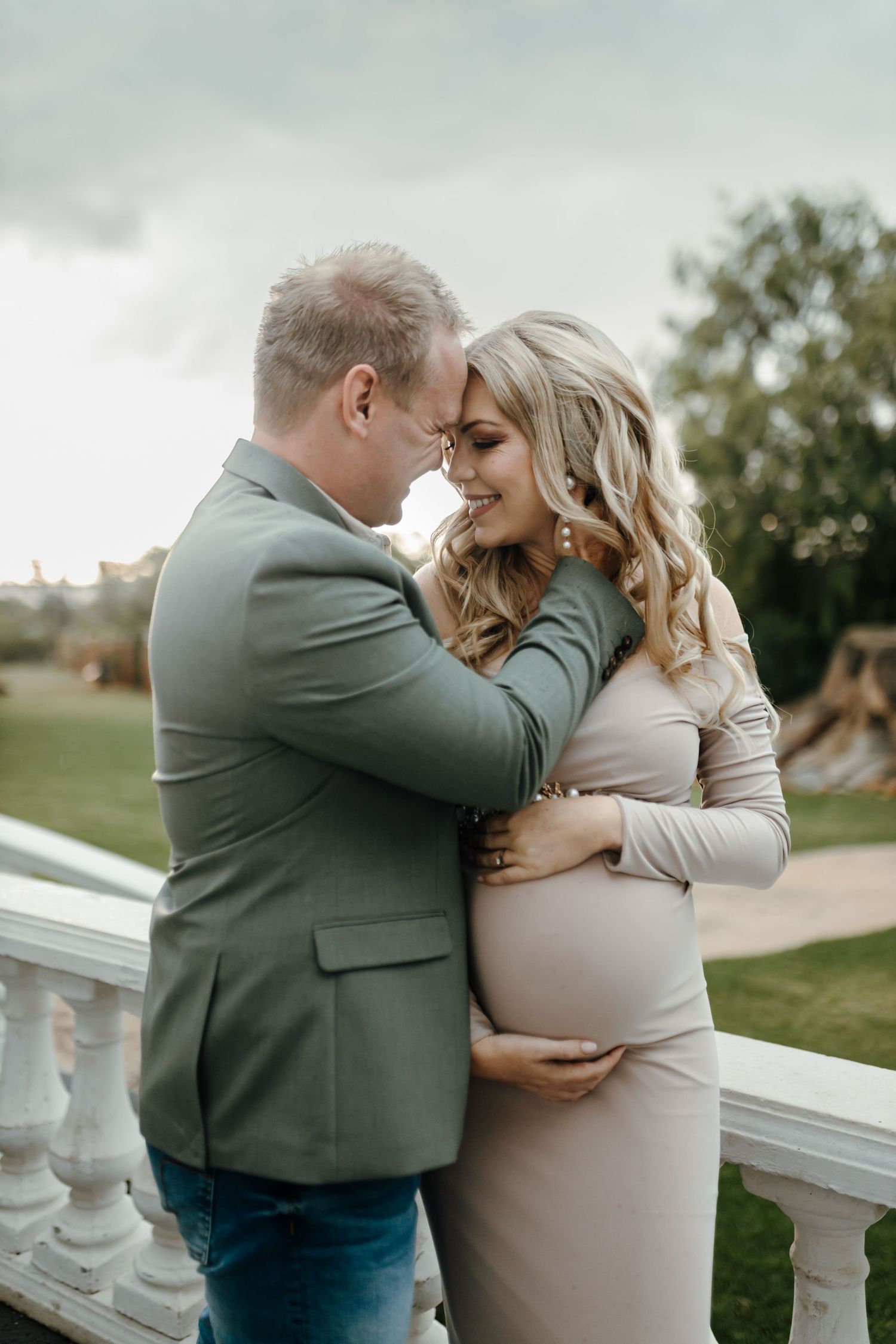 Romantic moment between a couple in soft beige and sage green tones near a white balustrade in a garden setting.
