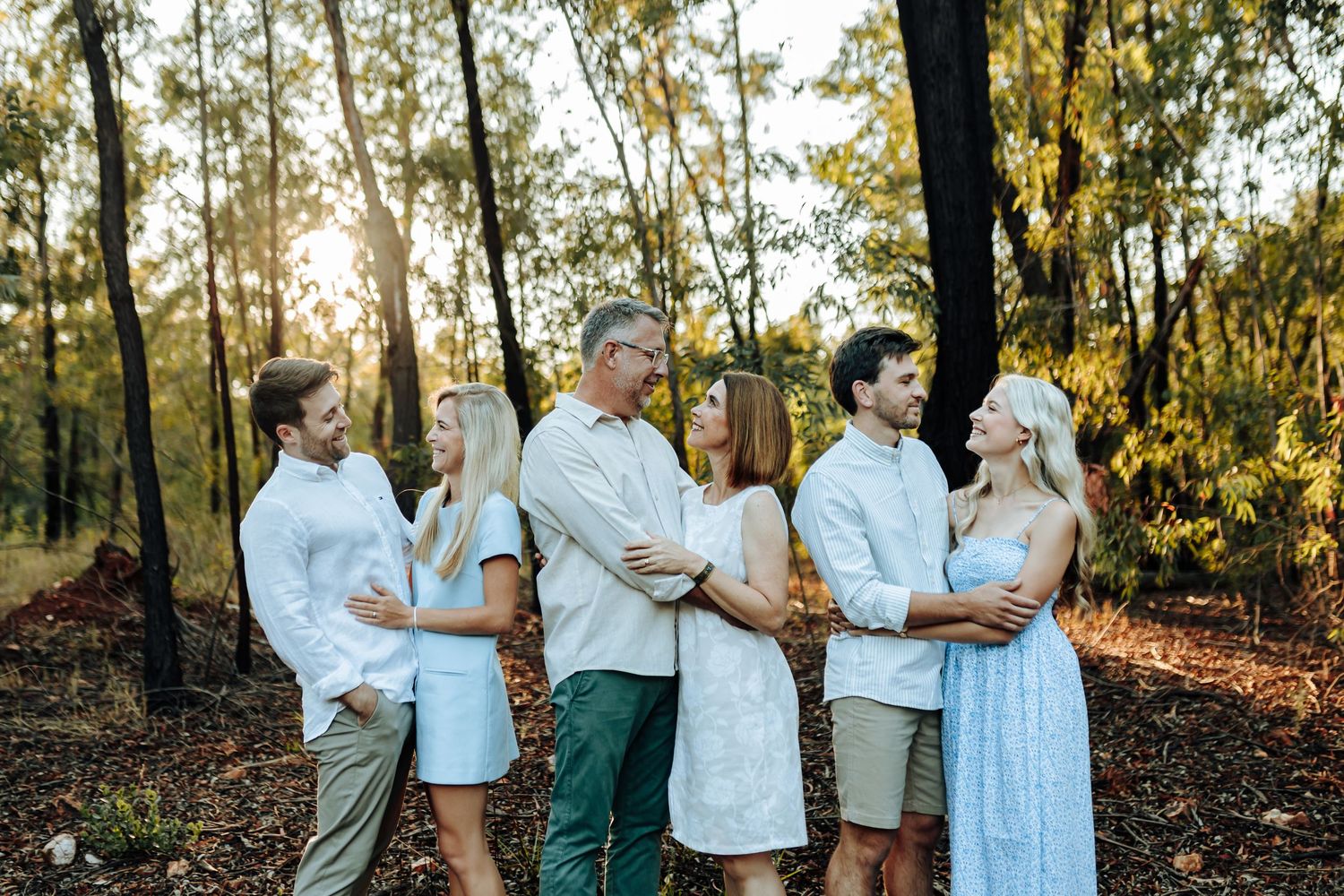 Family members in matching white and beige outfits pose together in a sunlit wooded area during golden hour.