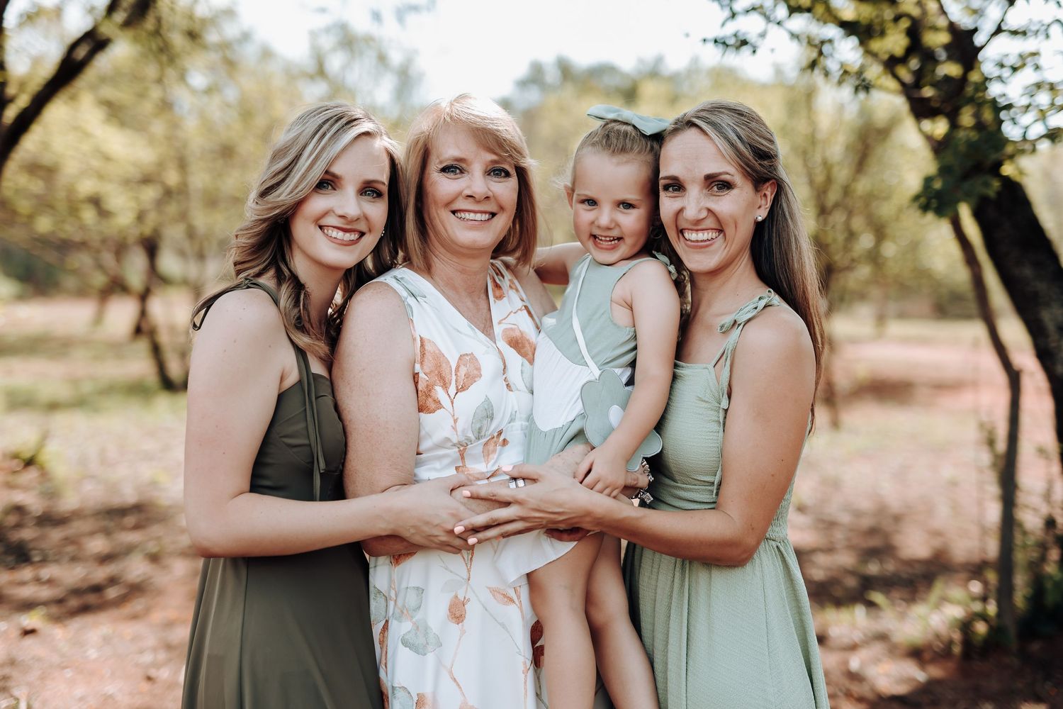 A group poses together in sage green dresses and white floral dress against a natural outdoor backdrop.