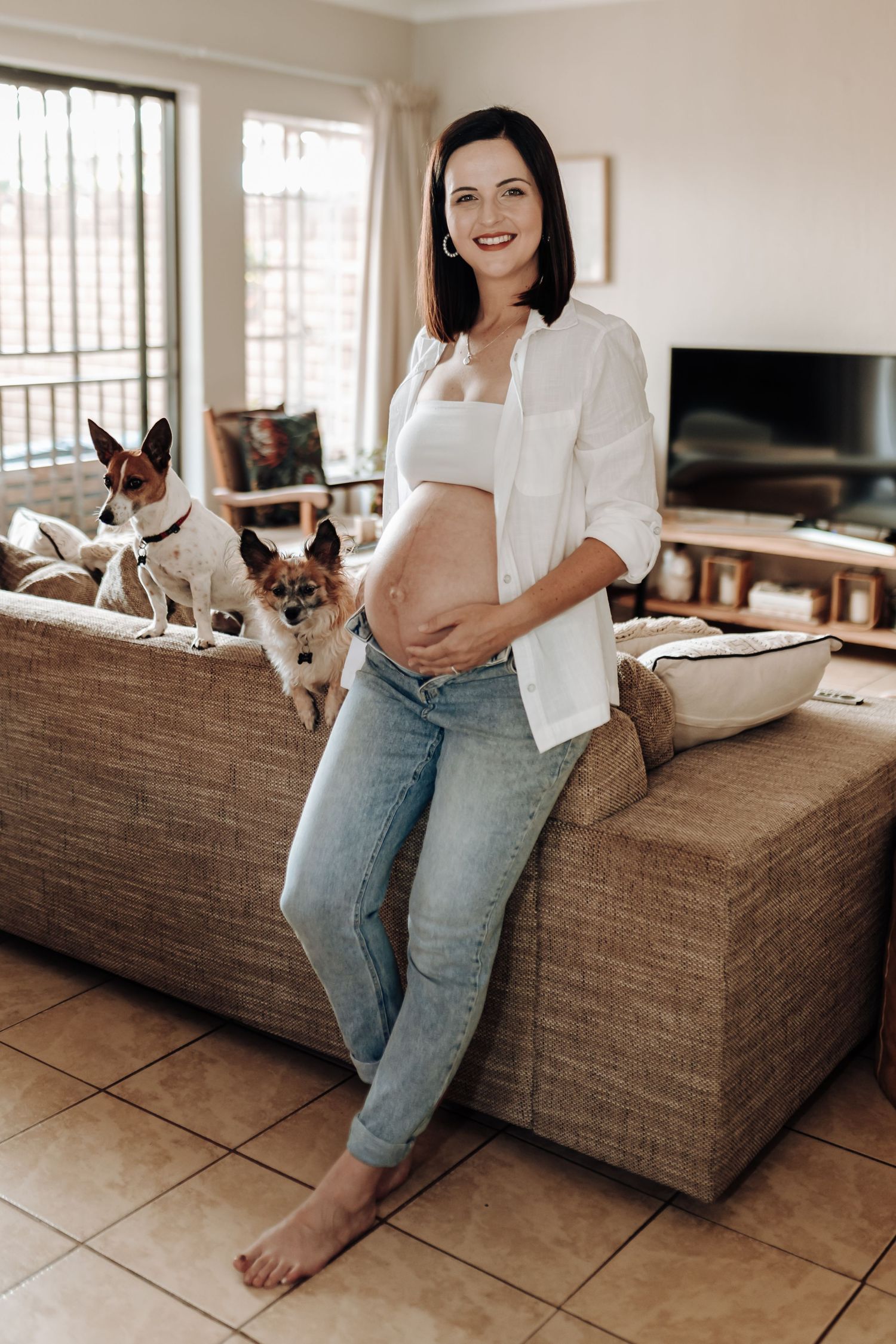 A woman in denim sits on a brown couch with a small chihuahua dog nearby.