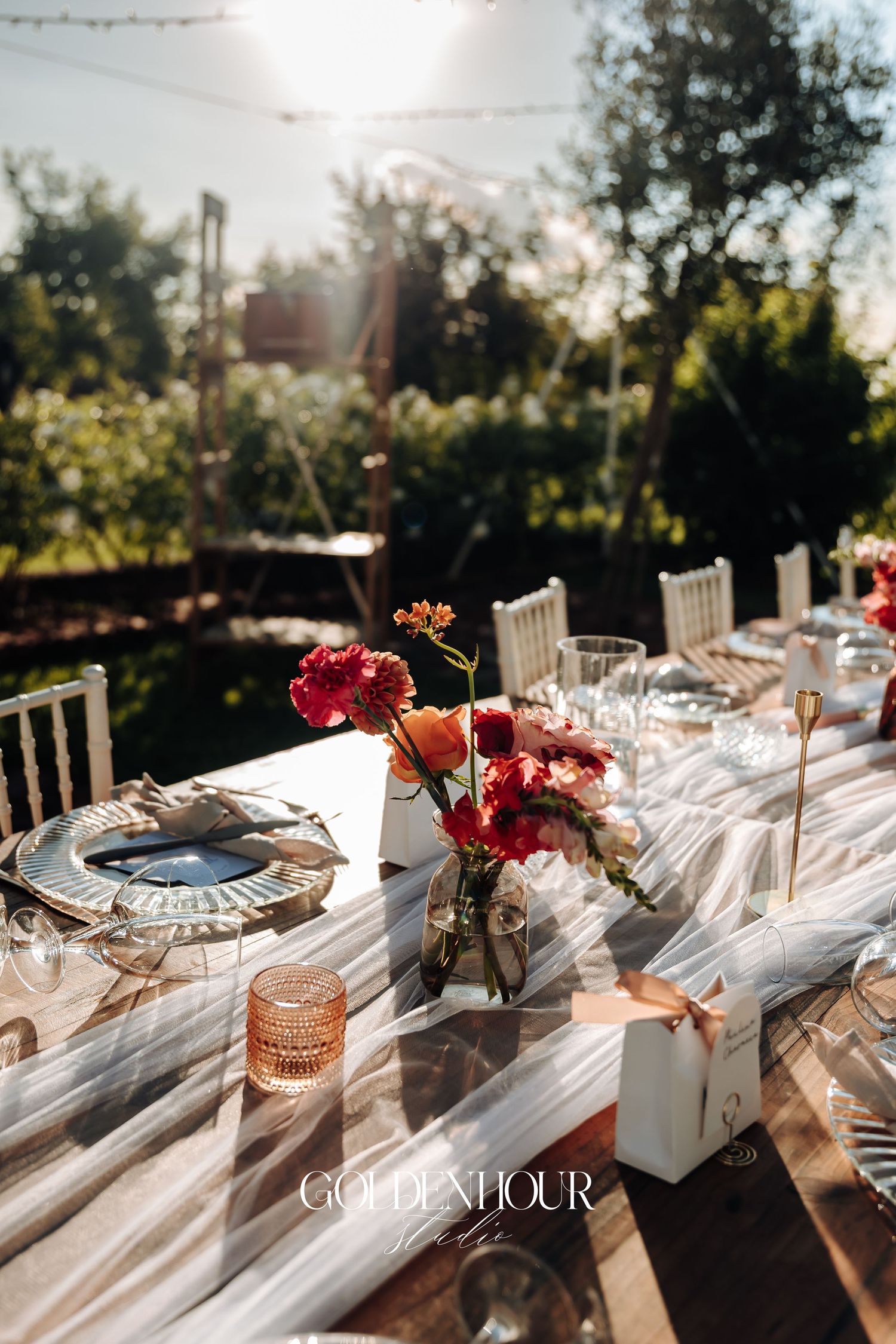 Elegant outdoor wedding reception table with white linens, red flowers, and glassware set against a sunset backdrop.