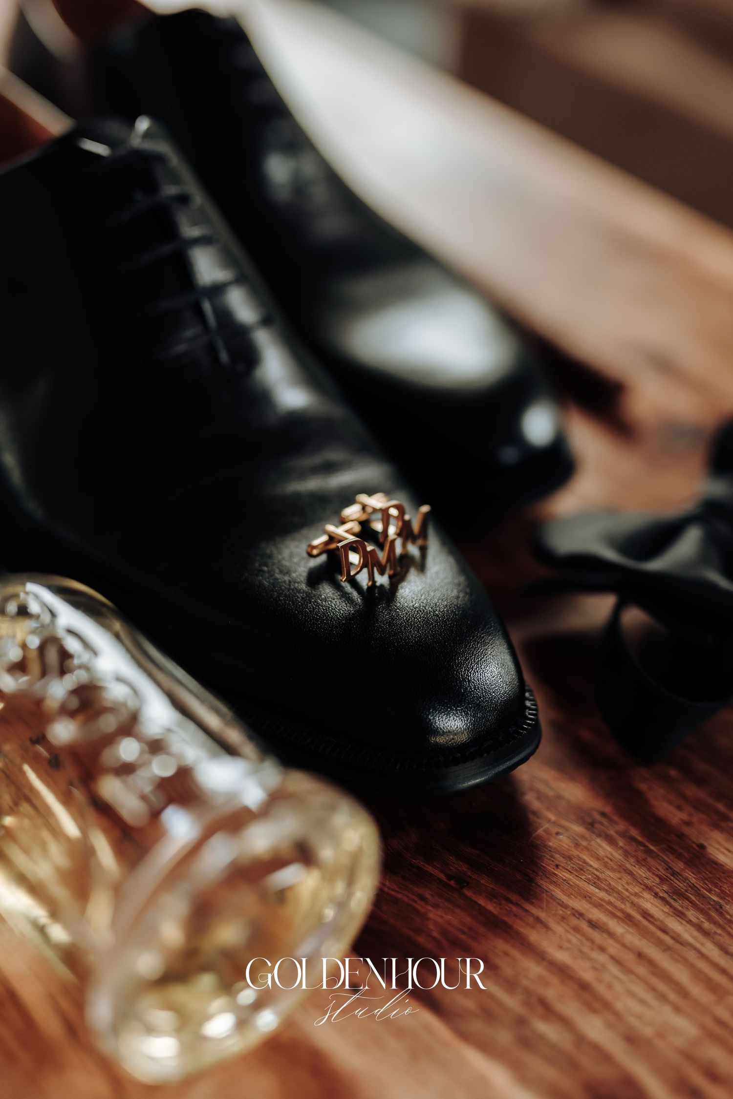Elegant black leather dress shoes with golden cufflinks sit beside a glass bottle on a wooden surface.