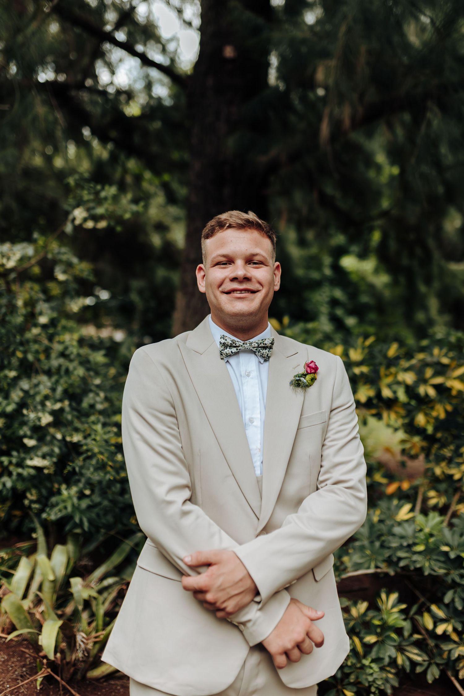 A wedding party member in a light beige suit with bow tie poses in a garden setting.