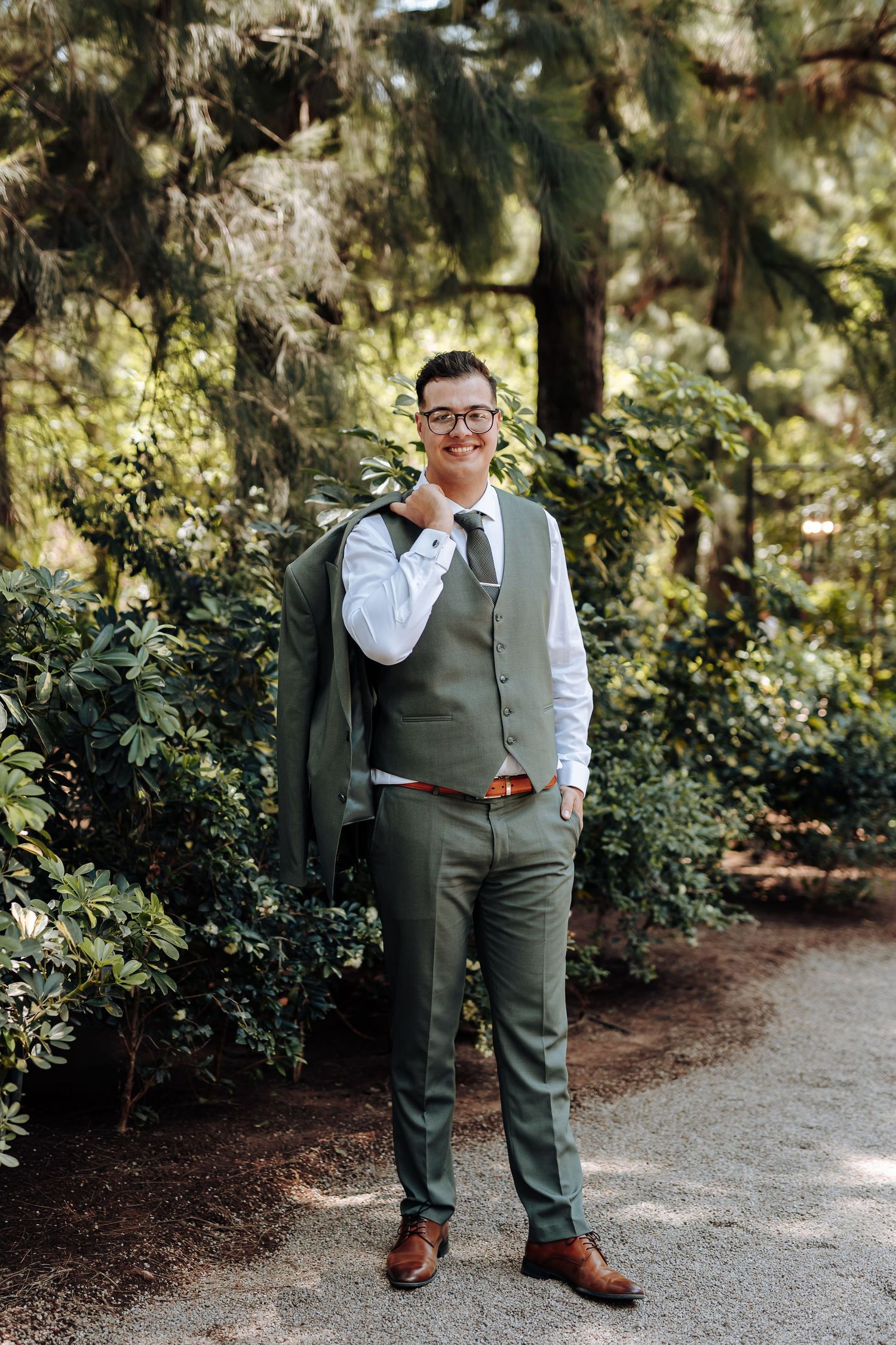 Person in formal attire poses casually on a garden path surrounded by lush greenery and trees.