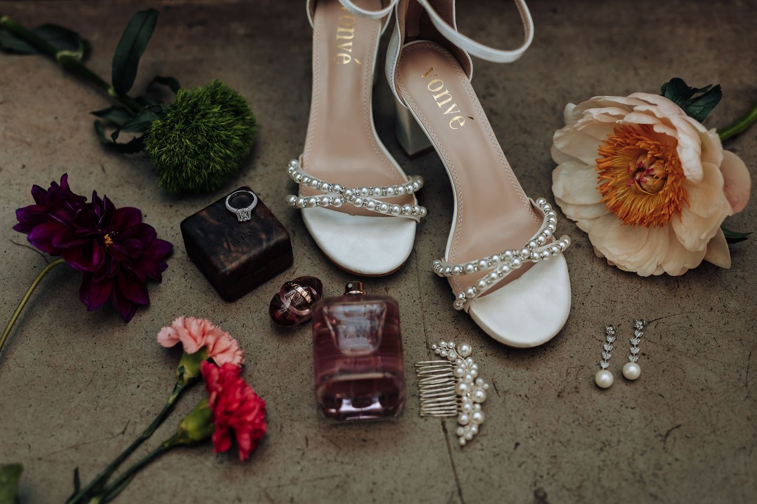 Elegant white bridal shoes surrounded by delicate flowers and jewelry accessories on a rustic concrete surface.