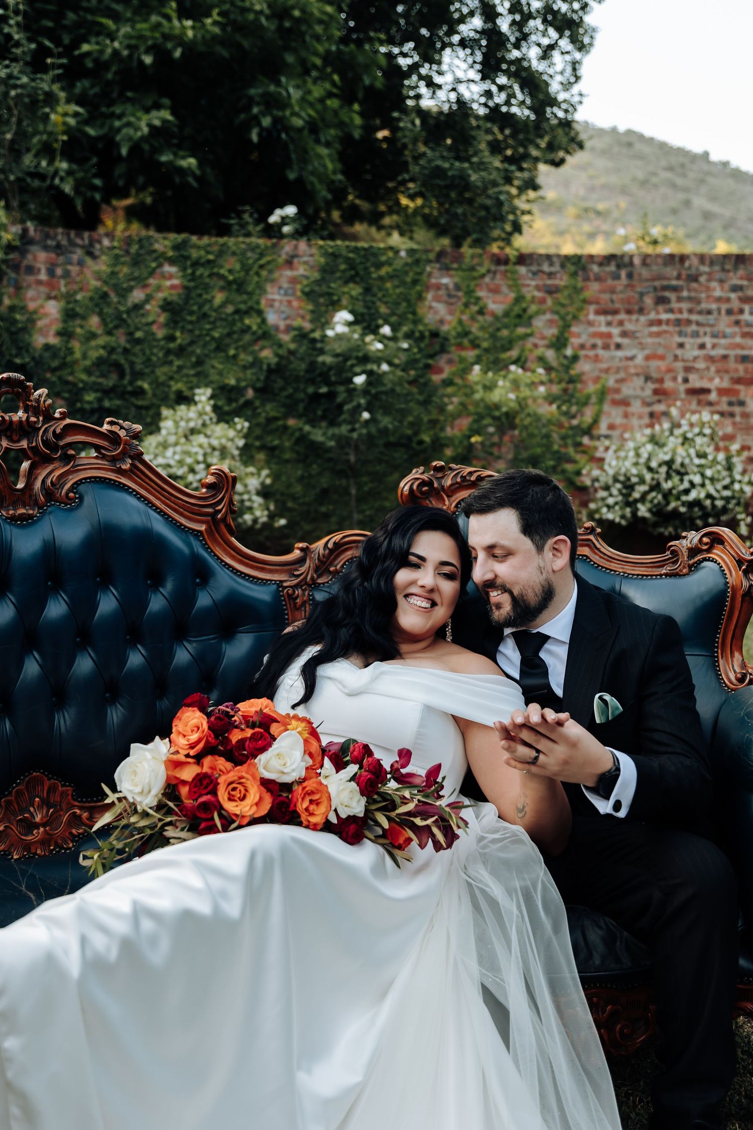 Newlyweds pose on a vintage blue tufted couch with orange floral arrangements in an outdoor garden setting.