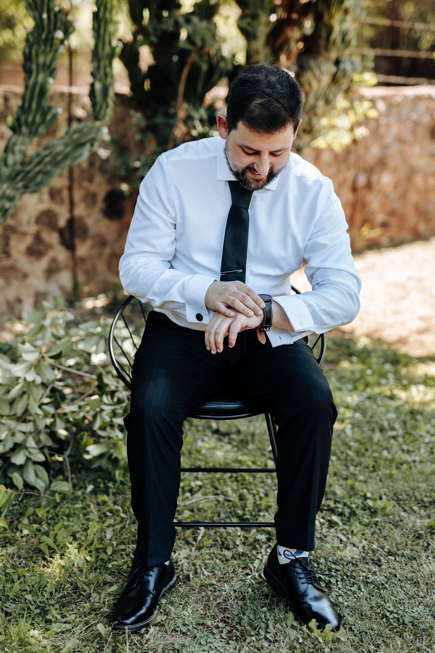 Person in formal attire sitting on chair while tying dress shoes in garden setting.