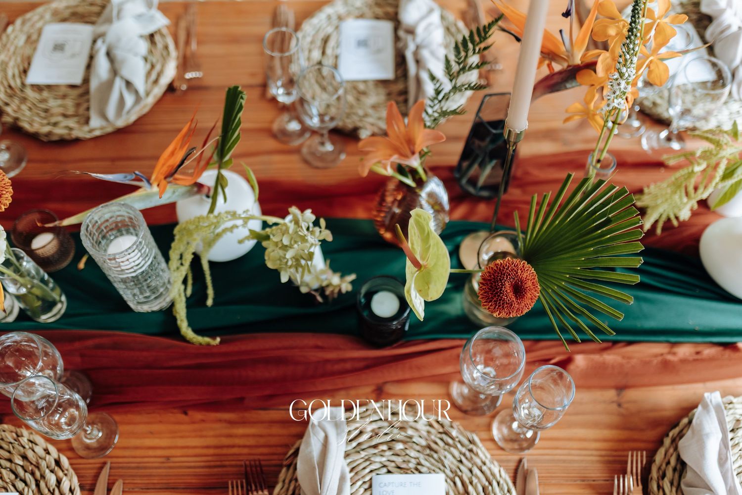 Elegant table setting with tropical greenery, gold accents, and white candles on a wooden surface for a wedding reception.