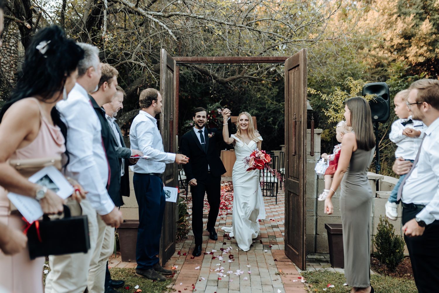 Wedding guests line pathway to greet newlyweds walking through wooden garden archway.