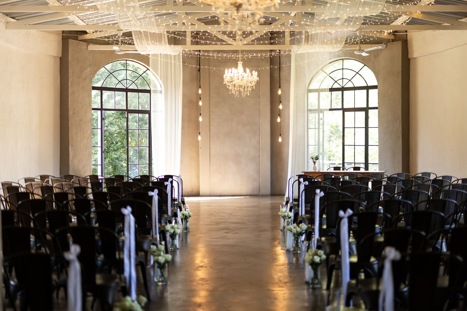 An elegant wedding ceremony venue with arched windows, crystal chandelier, and rows of dark chairs in a bright white room.