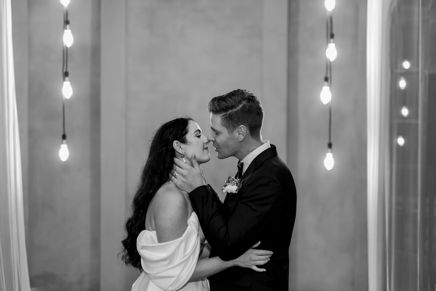 A romantic black and white wedding portrait with hanging Edison bulbs creating an intimate atmosphere.