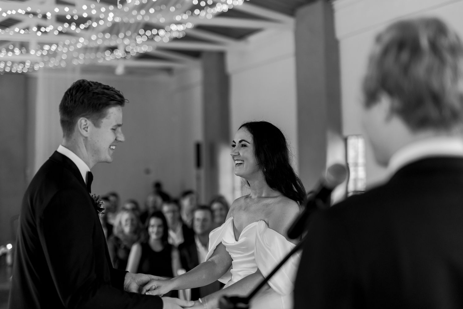 A couple exchanging wedding vows in an elegant venue with chandeliers and guests looking on.