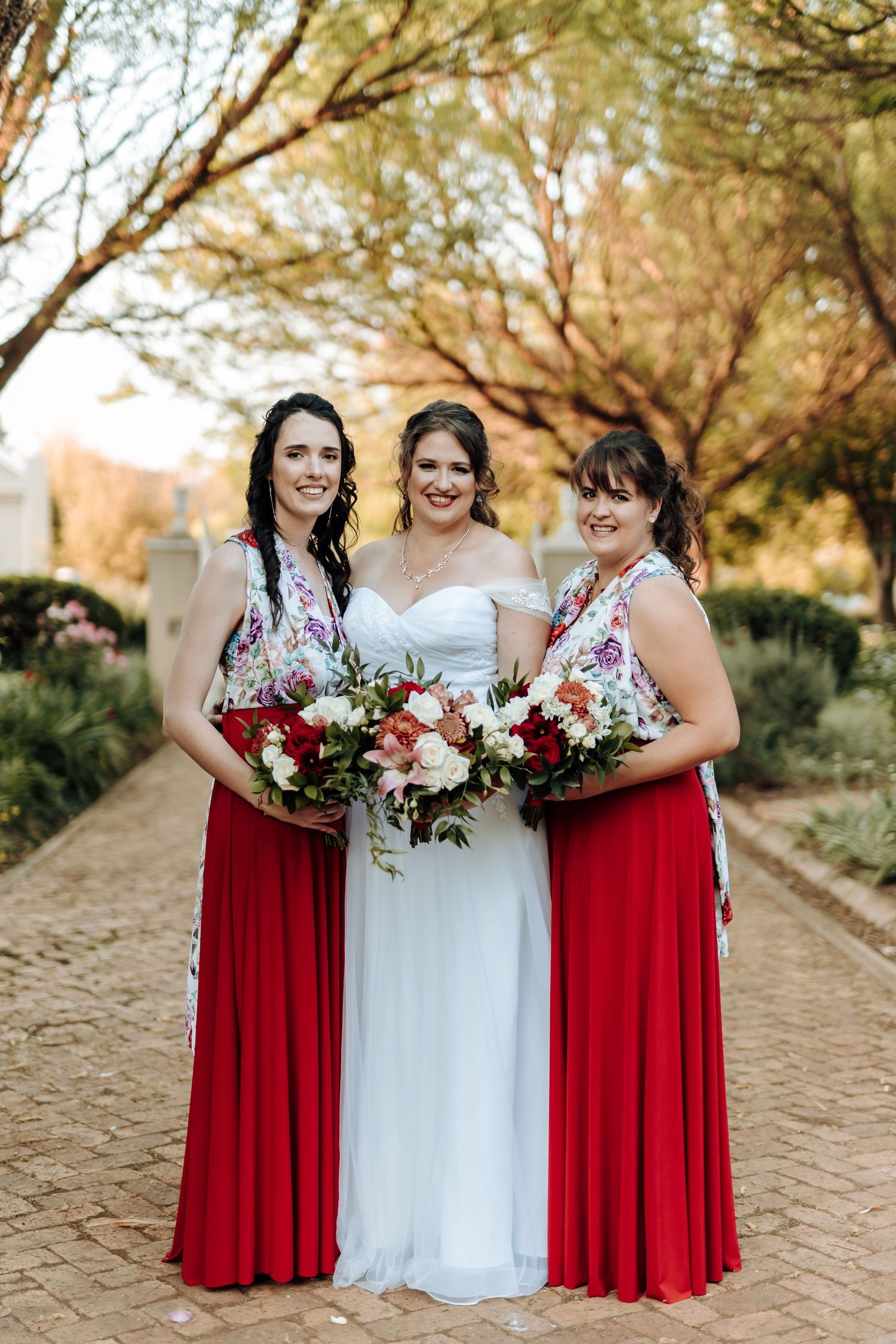 Three women in coordinated red and white attire smile while holding floral bouquets on a brick path.