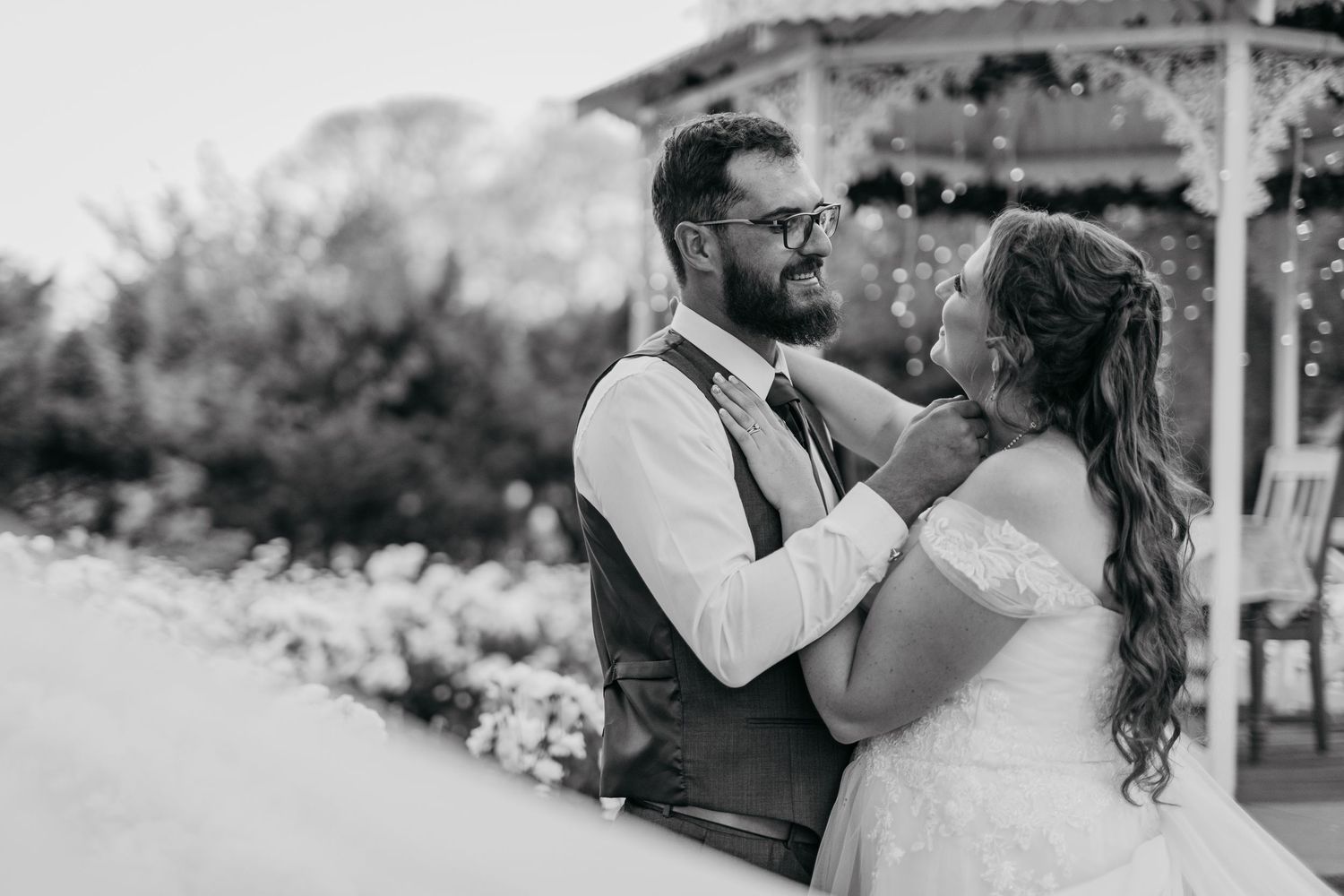 Black and white photo of a couple sharing a romantic moment by a garden gazebo.