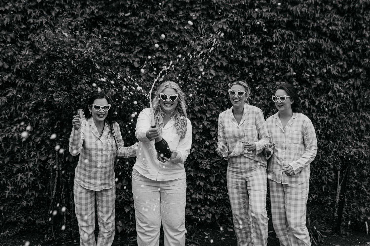 Black and white photo of friends celebrating with champagne against a hedge background.