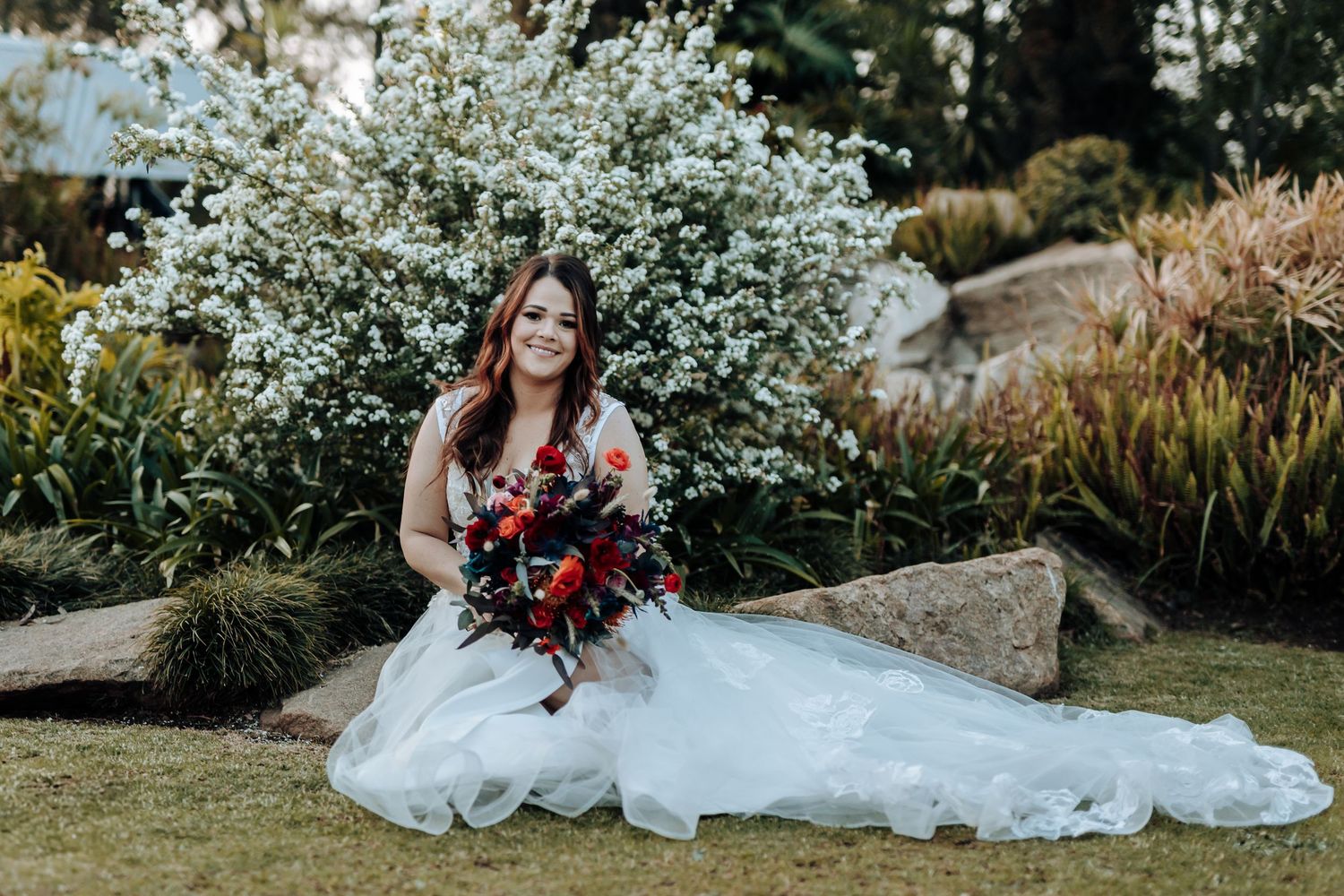 Bride sits on garden rocks holding red bouquet against flowering bush backdrop.