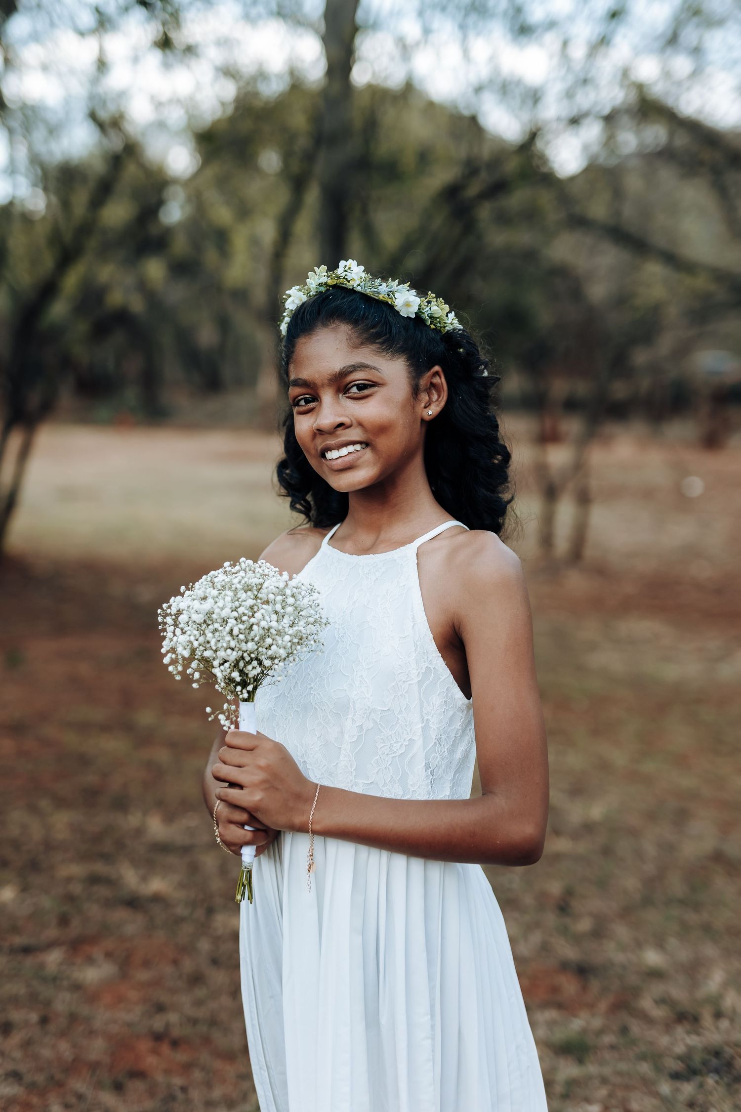 Girl in white dress holds baby's breath bouquet wearing flower crown in outdoor setting.