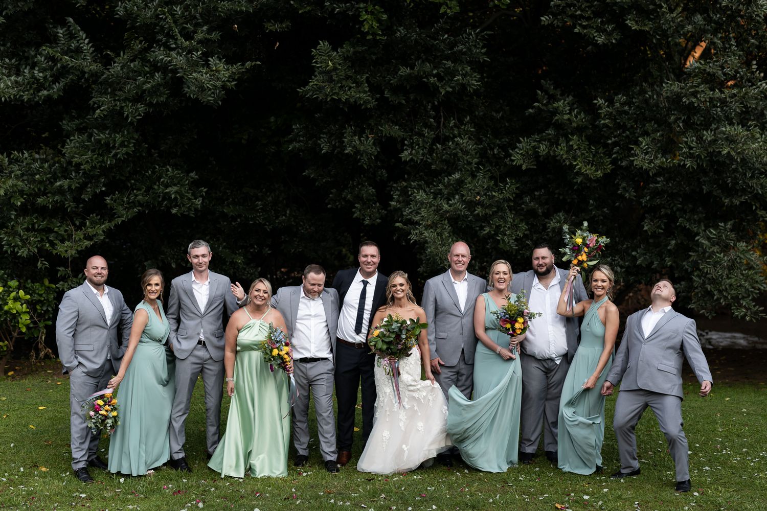 Wedding party in grey suits and mint dresses posed casually on lawn.