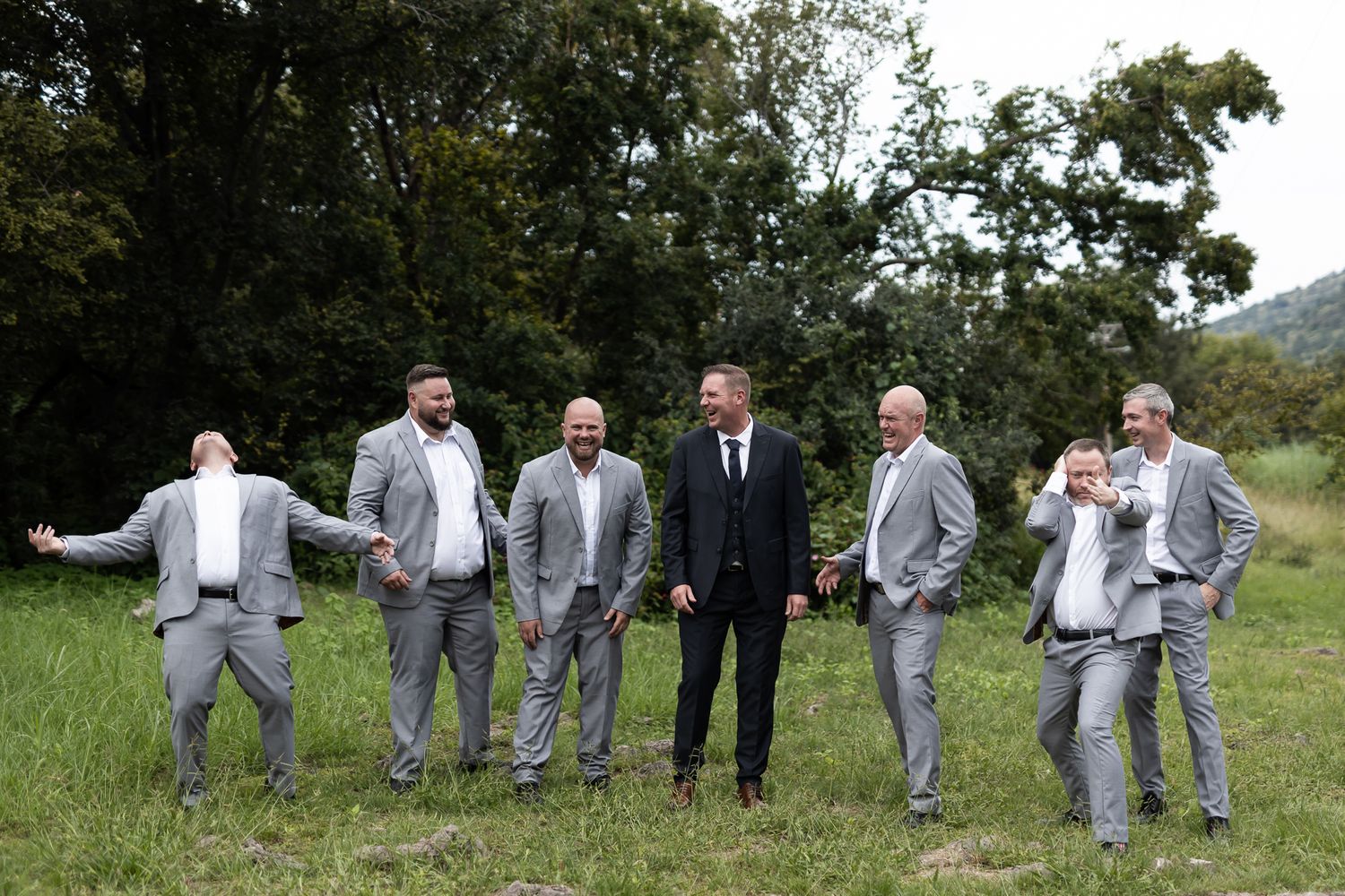 Groomsmen in light gray suits strike playful poses alongside one in black suit in an outdoor wedding photo.