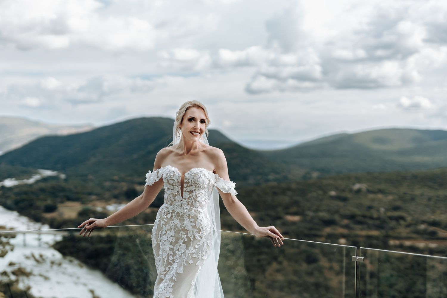Bride in off-shoulder lace gown poses against glass railing with mountain landscape behind.