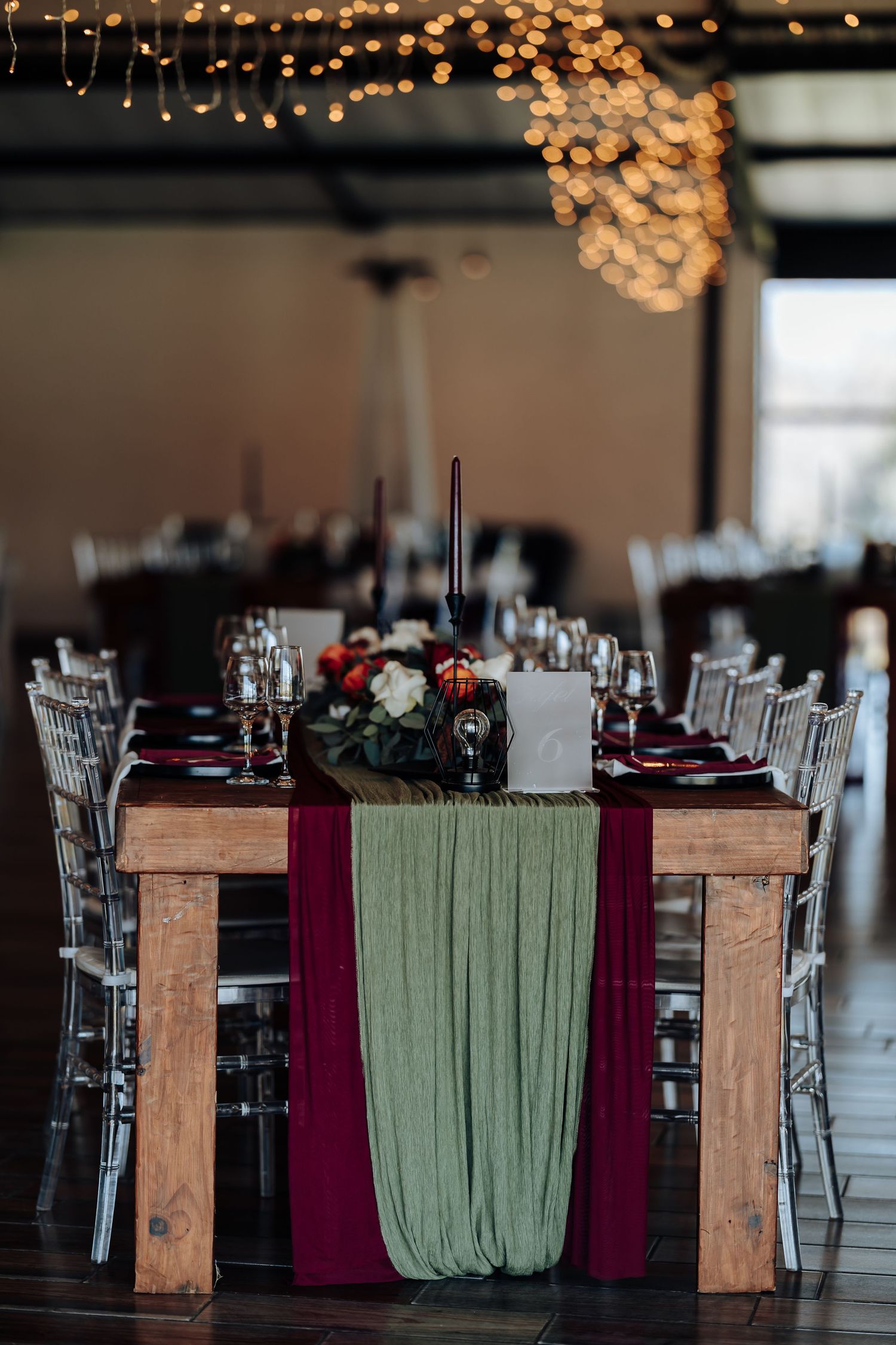 Elegant wedding reception table with burgundy and sage green runners, floral centerpiece, dark candles, and fairy lights overhead.