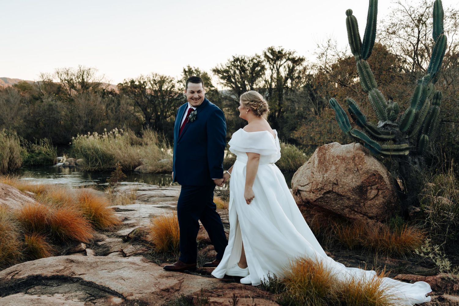 Bride in white off-shoulder gown and groom in navy suit stand on desert rocks near cacti and a pond at sunset.