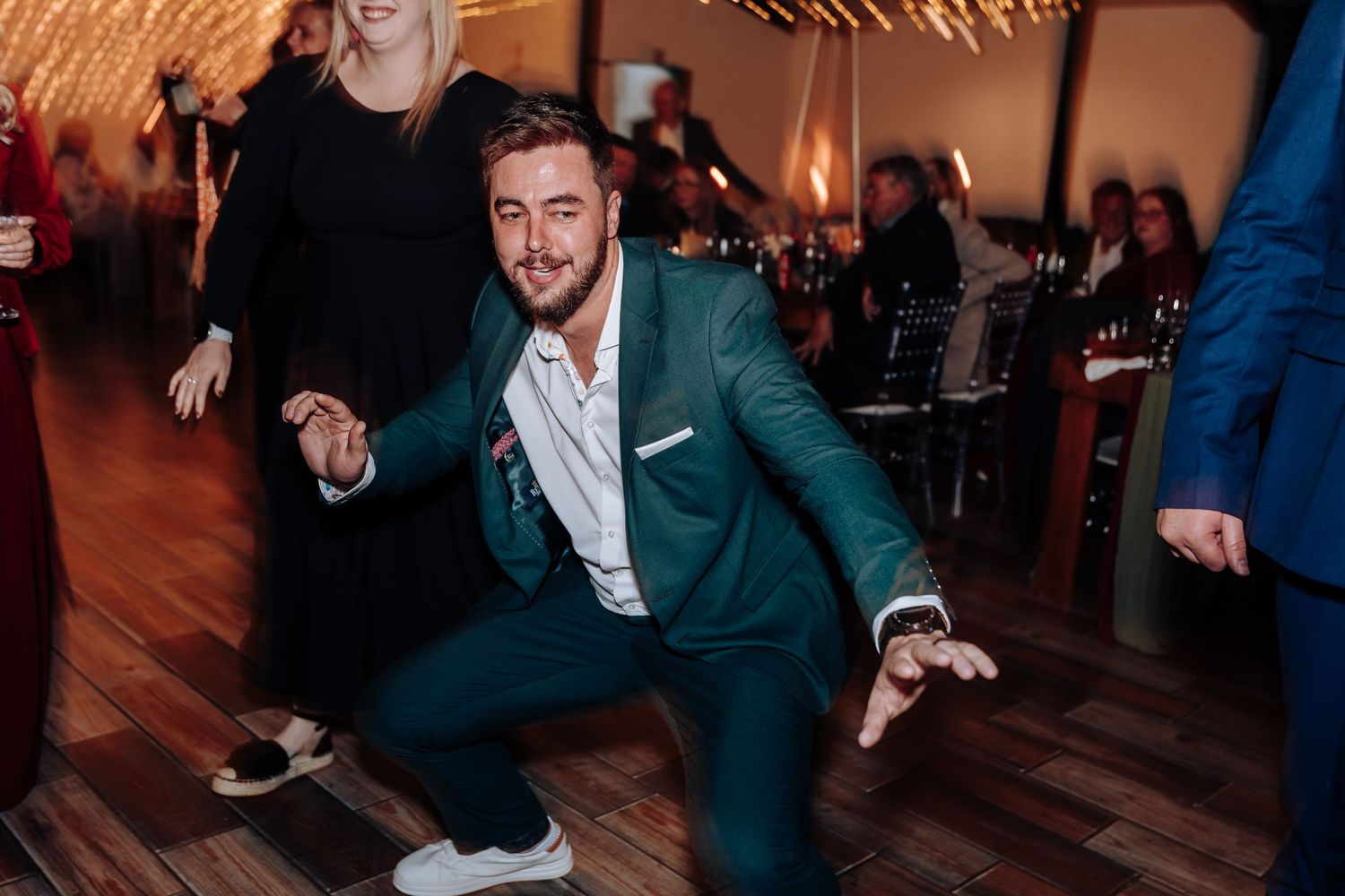 Man in green suit dancing energetically at a wedding reception on a wooden dance floor.