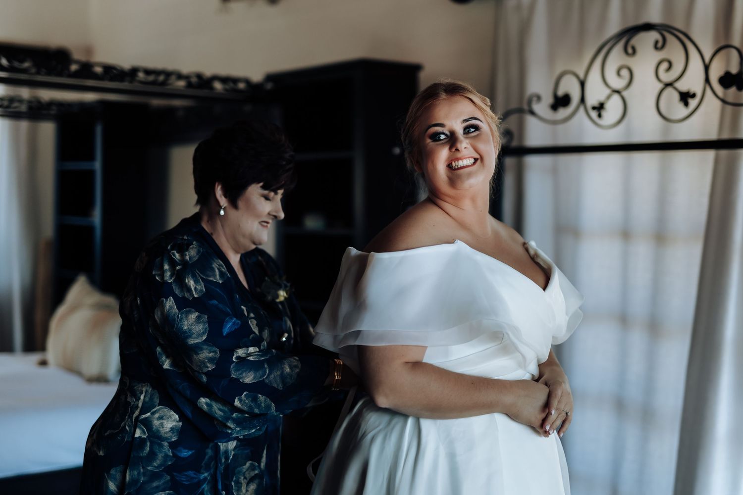 A bride in a white off-shoulder gown smiles as a woman in floral dress helps fasten her wedding dress in a elegant room.