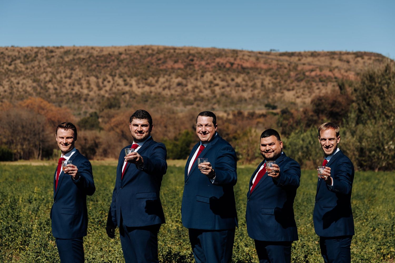 Five groomsmen in navy suits and red ties pose outdoors holding drinks with a scenic hill backdrop.